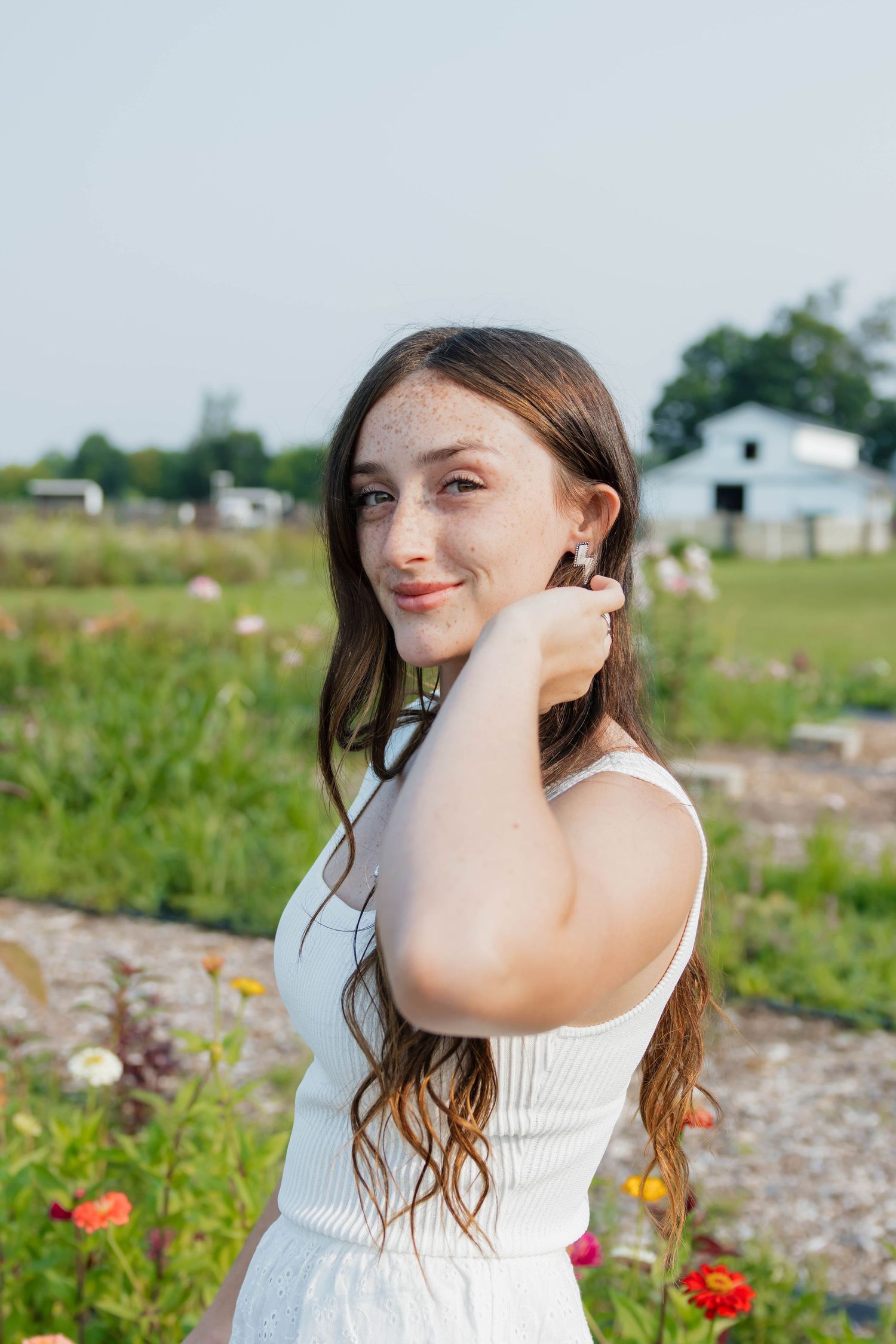 Smiling young woman with freckles in white dress touching hair in colorful outdoor flower garden.
