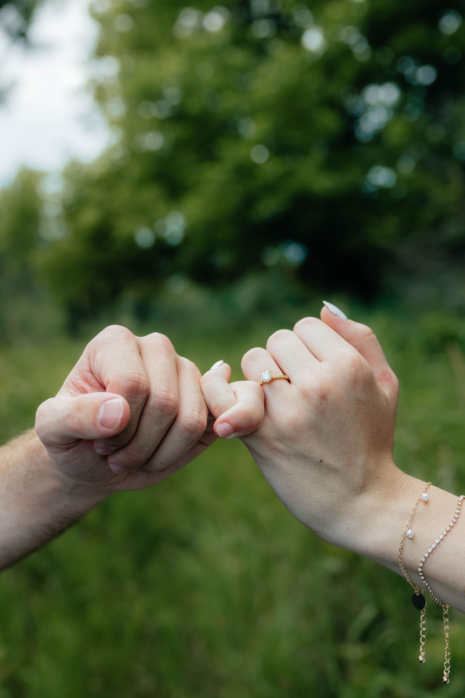 Close-up of couple making a pinky promise with engagement ring visible on woman's hand outdoors.