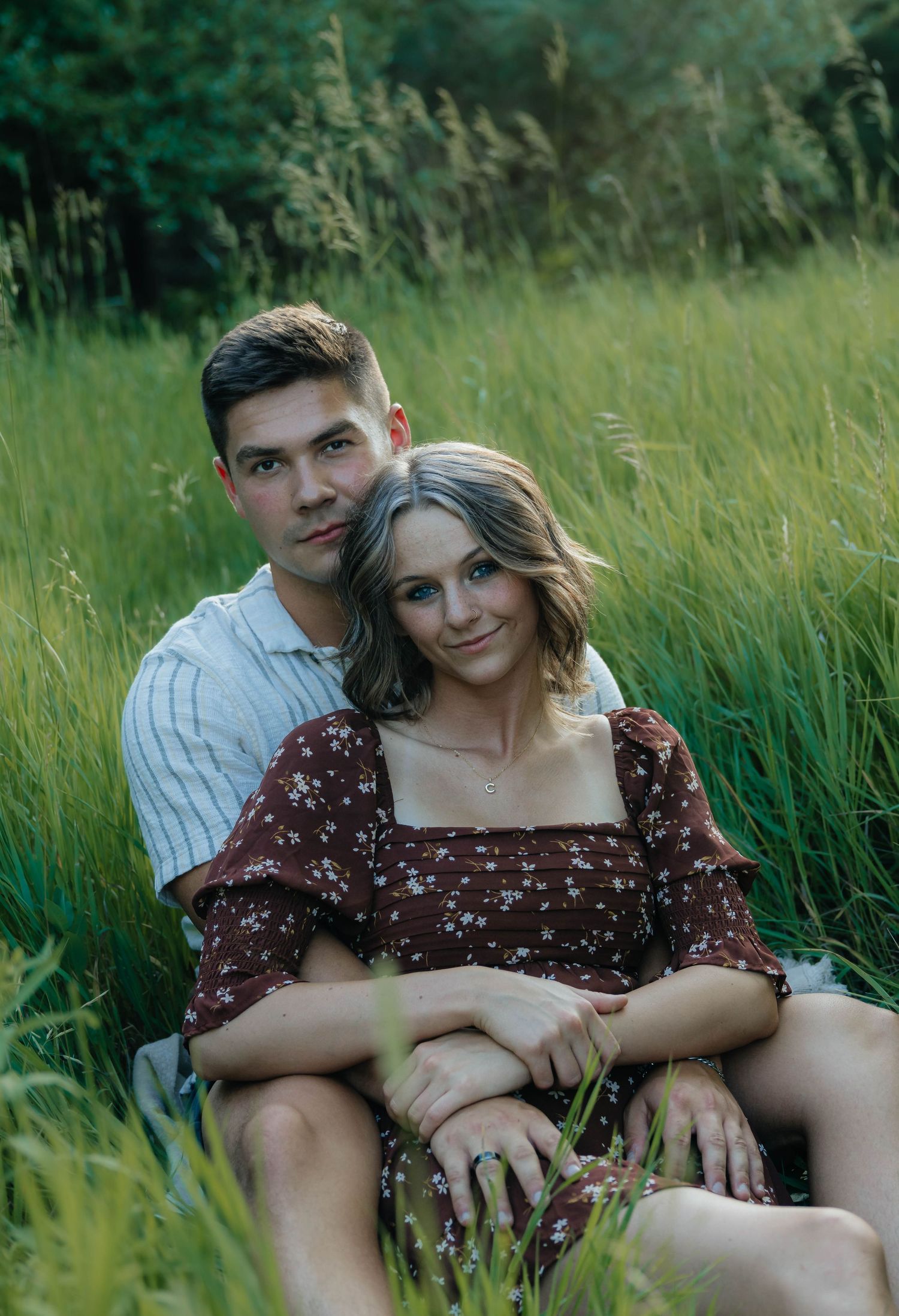 Couple sitting together in tall green grass field, woman leaning back against man, both smiling at camera.