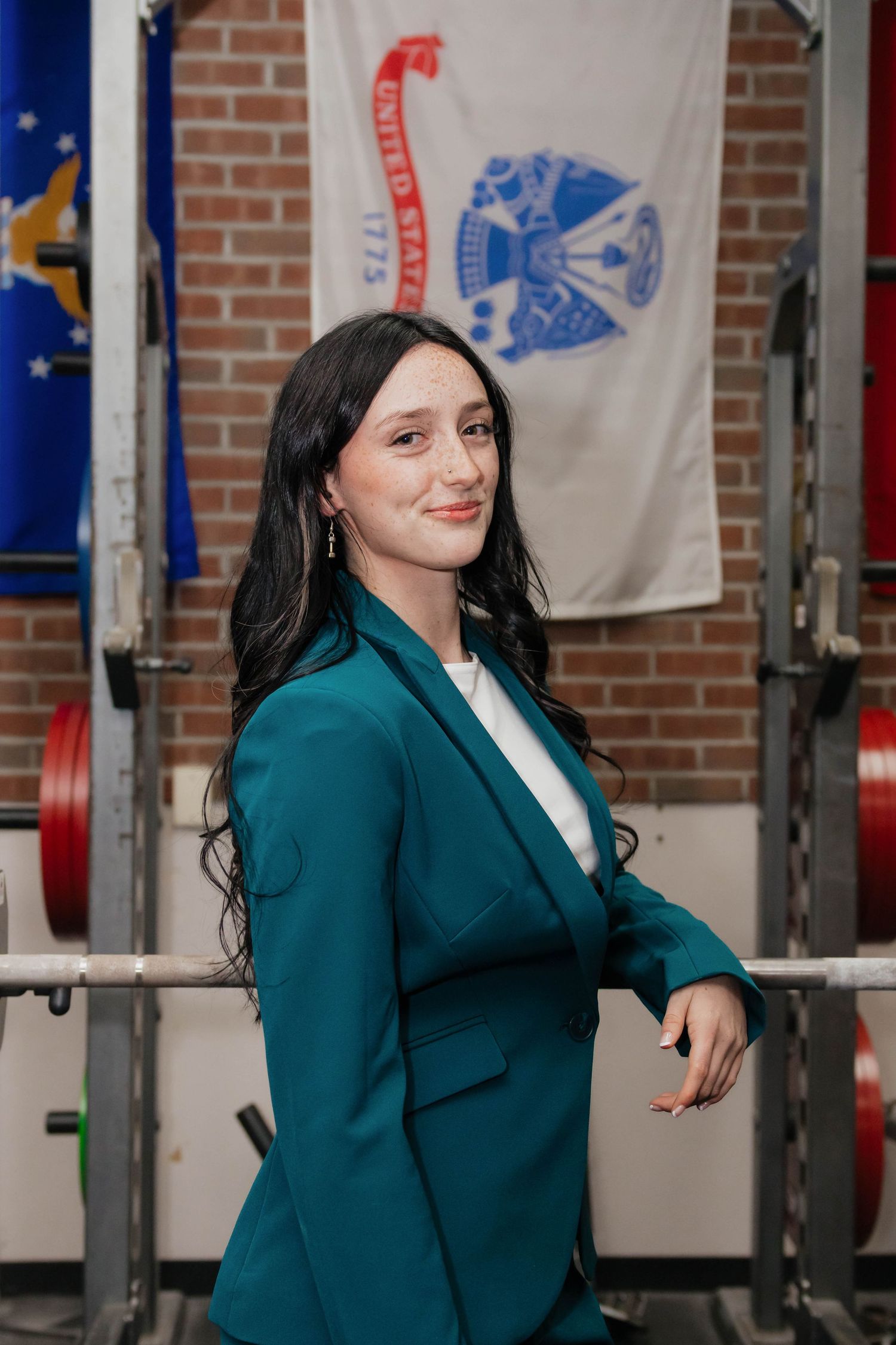 Woman in teal blazer posing in a gym with US Army flag displayed on brick wall behind her.