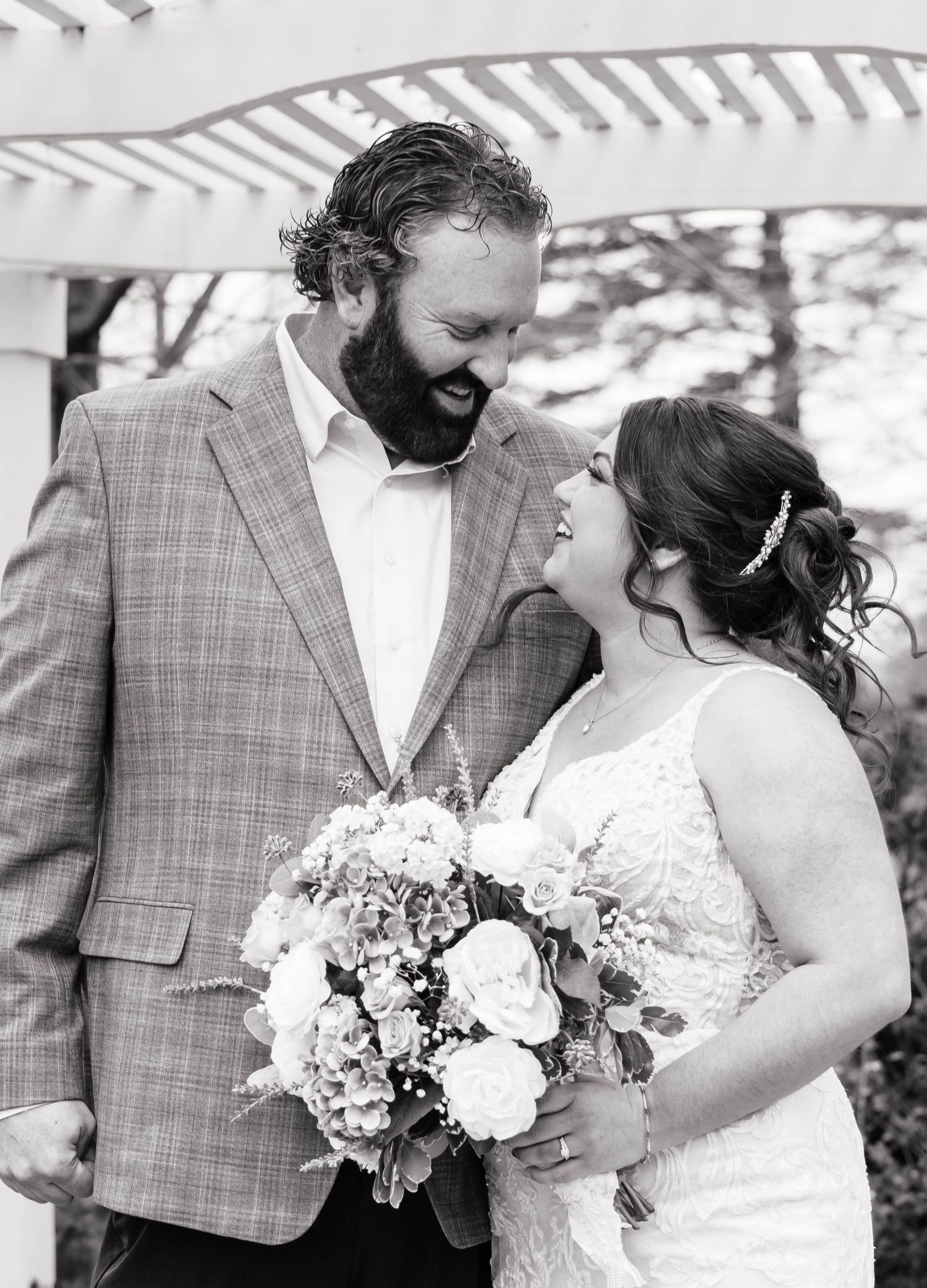 Black and white portrait of smiling bride and bearded groom sharing tender moment under garden arbor.