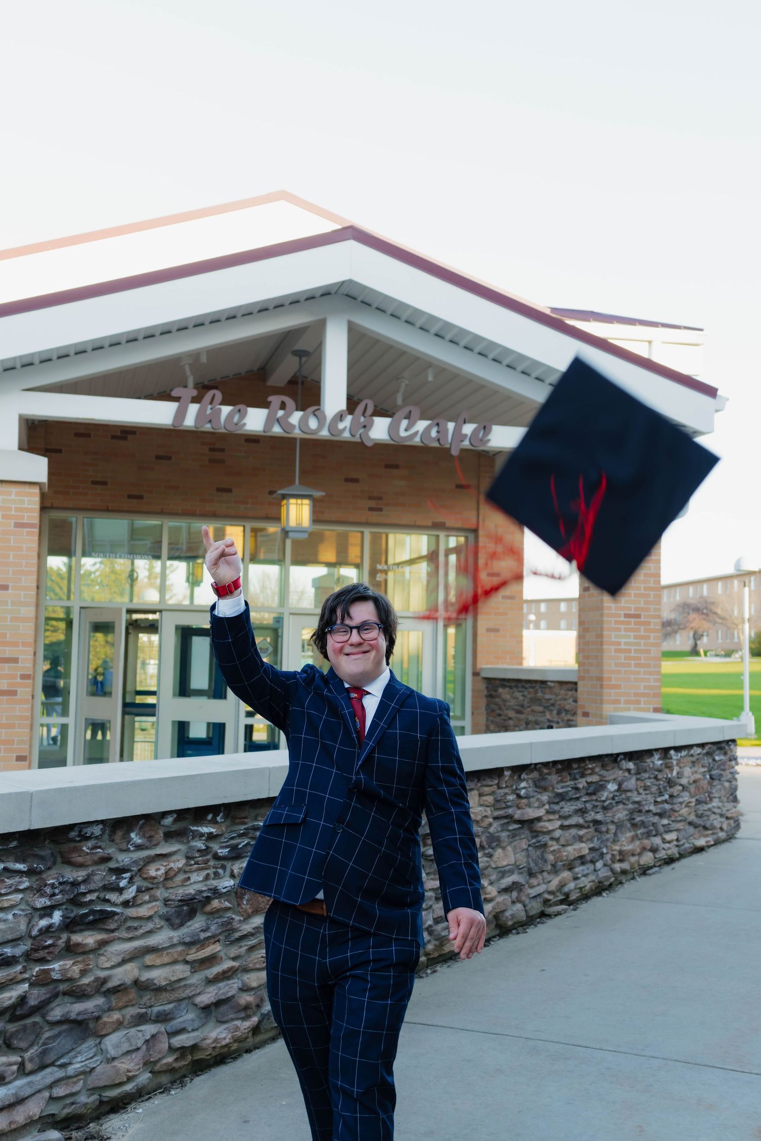 Excited male graduate in plaid suit tosses cap in air outside The Rock Cafe building.
