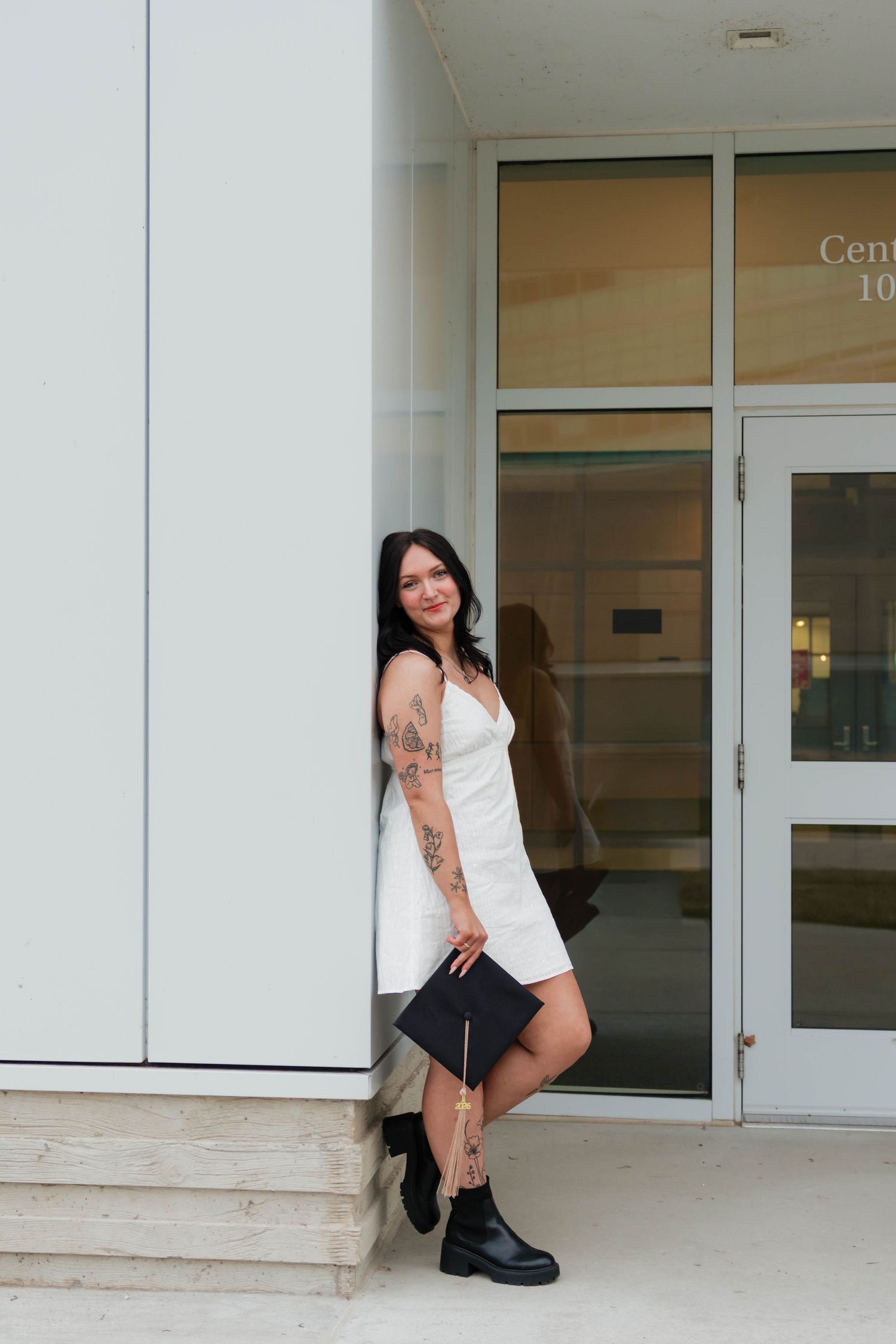 Tattooed graduate in white dress and black boots leaning against building entrance with cap.