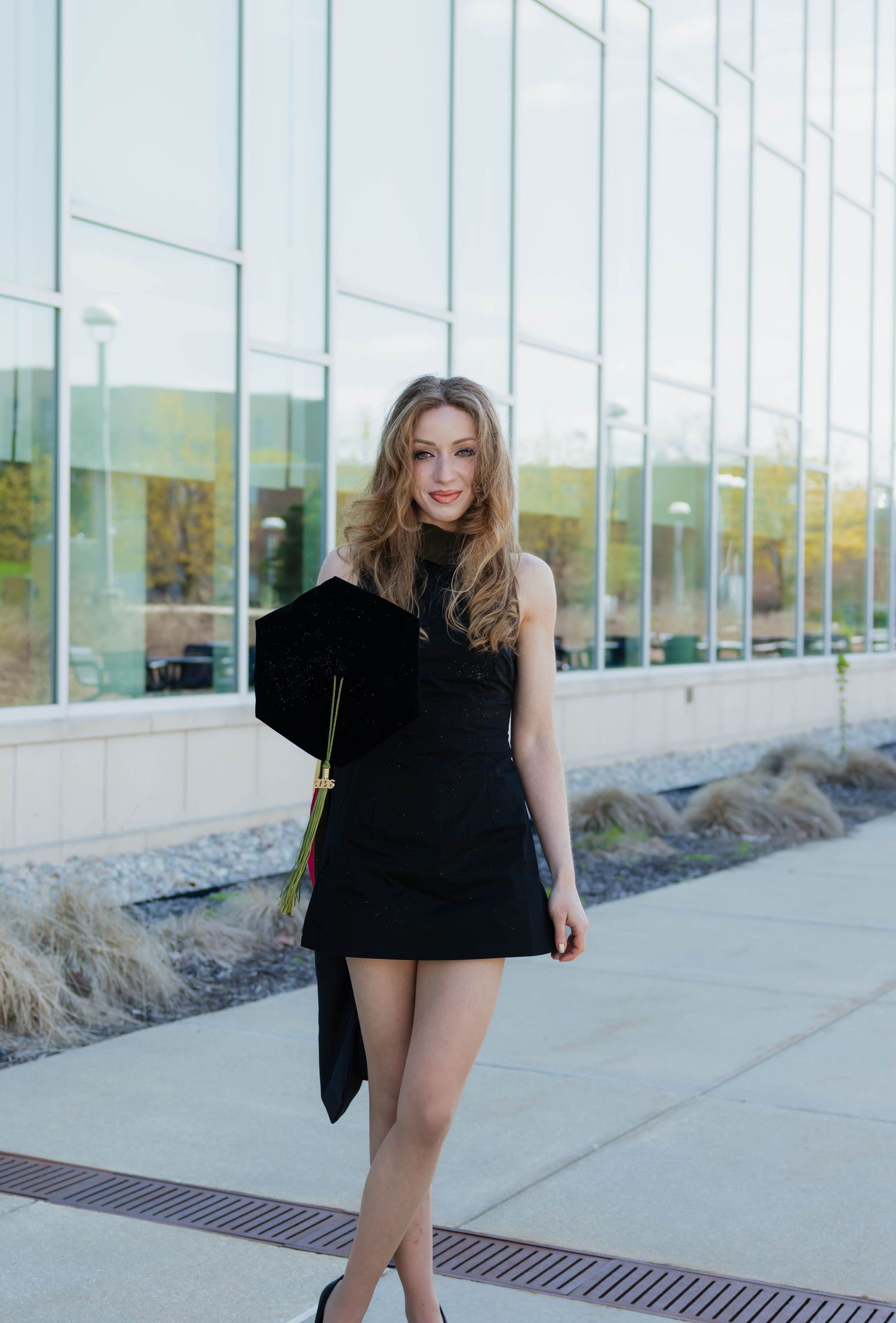 Elegant female graduate in black mini dress holding mortarboard outside glass-facade university building.
