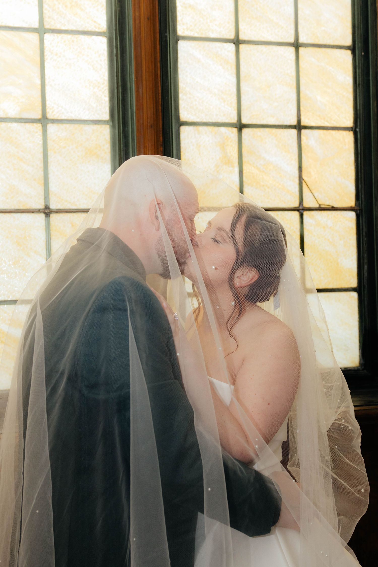 Bride and groom kiss under flowing veil in front of stained glass window.