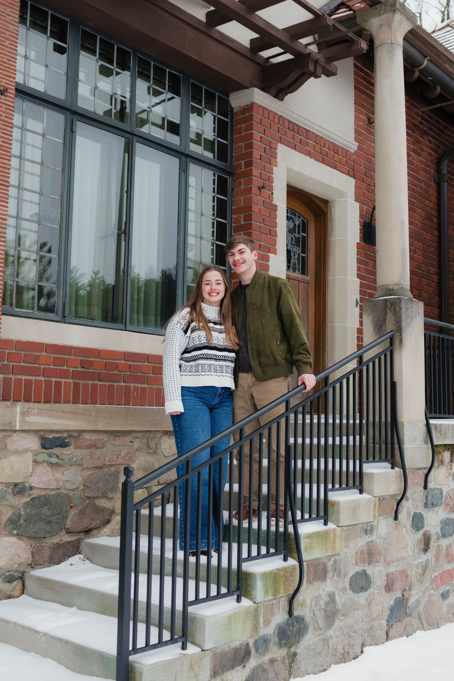 Smiling couple standing on snow-dusted steps outside a charming brick building in winter.
