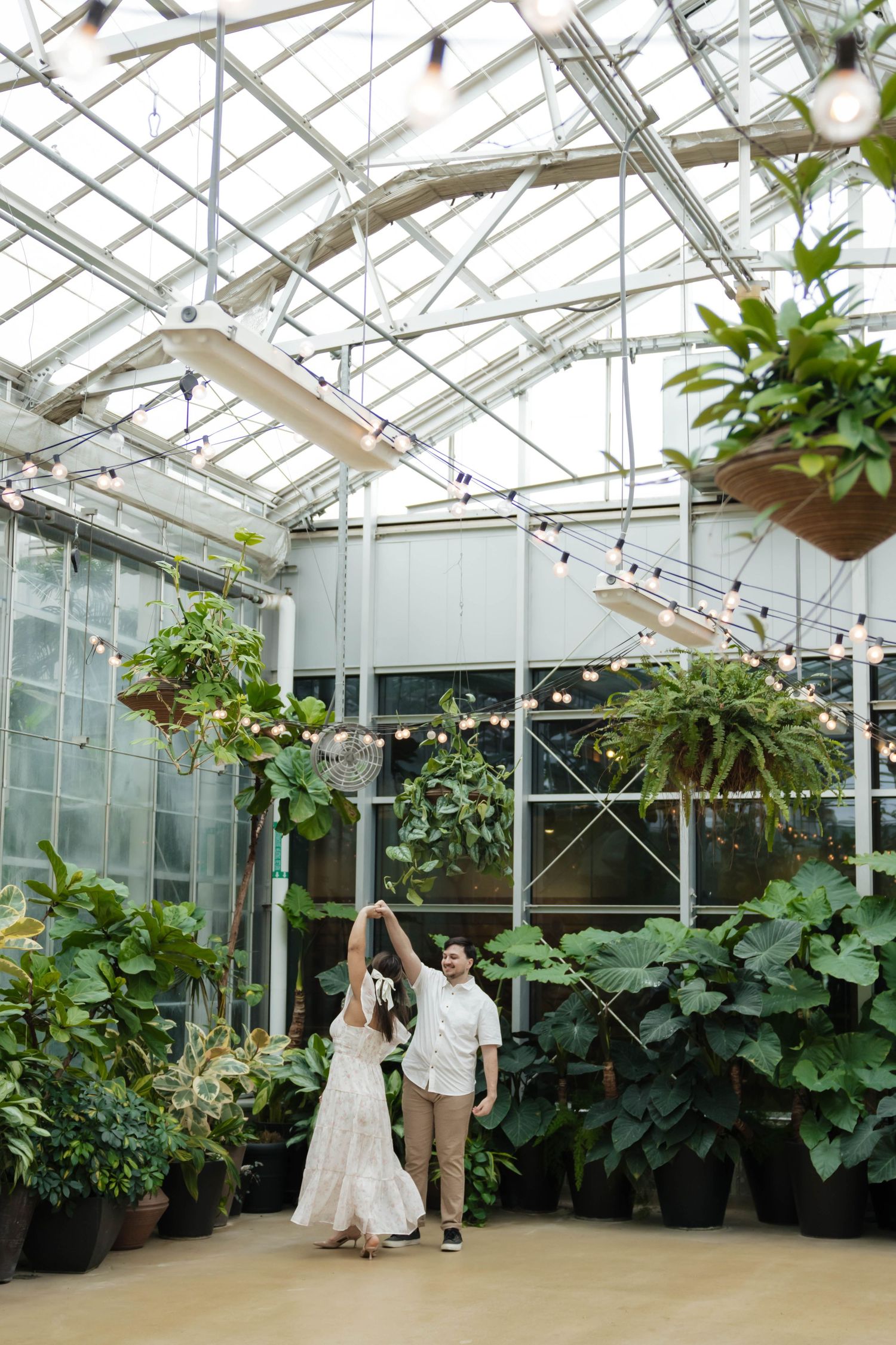 Couple dancing and spinning in a glass greenhouse filled with hanging plants and string lights.