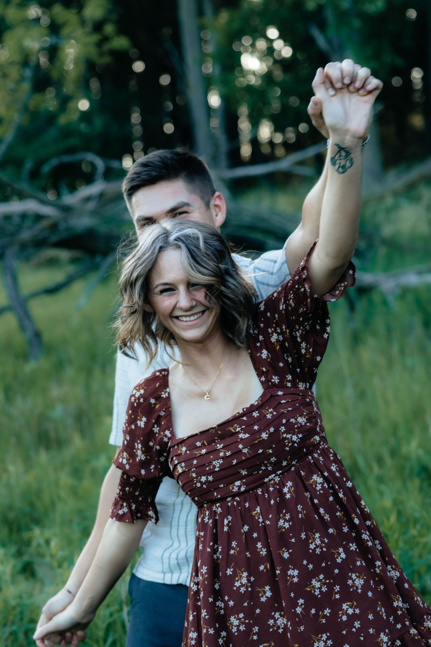 Joyful woman in burgundy floral dress laughing as partner spins her in a forest clearing.