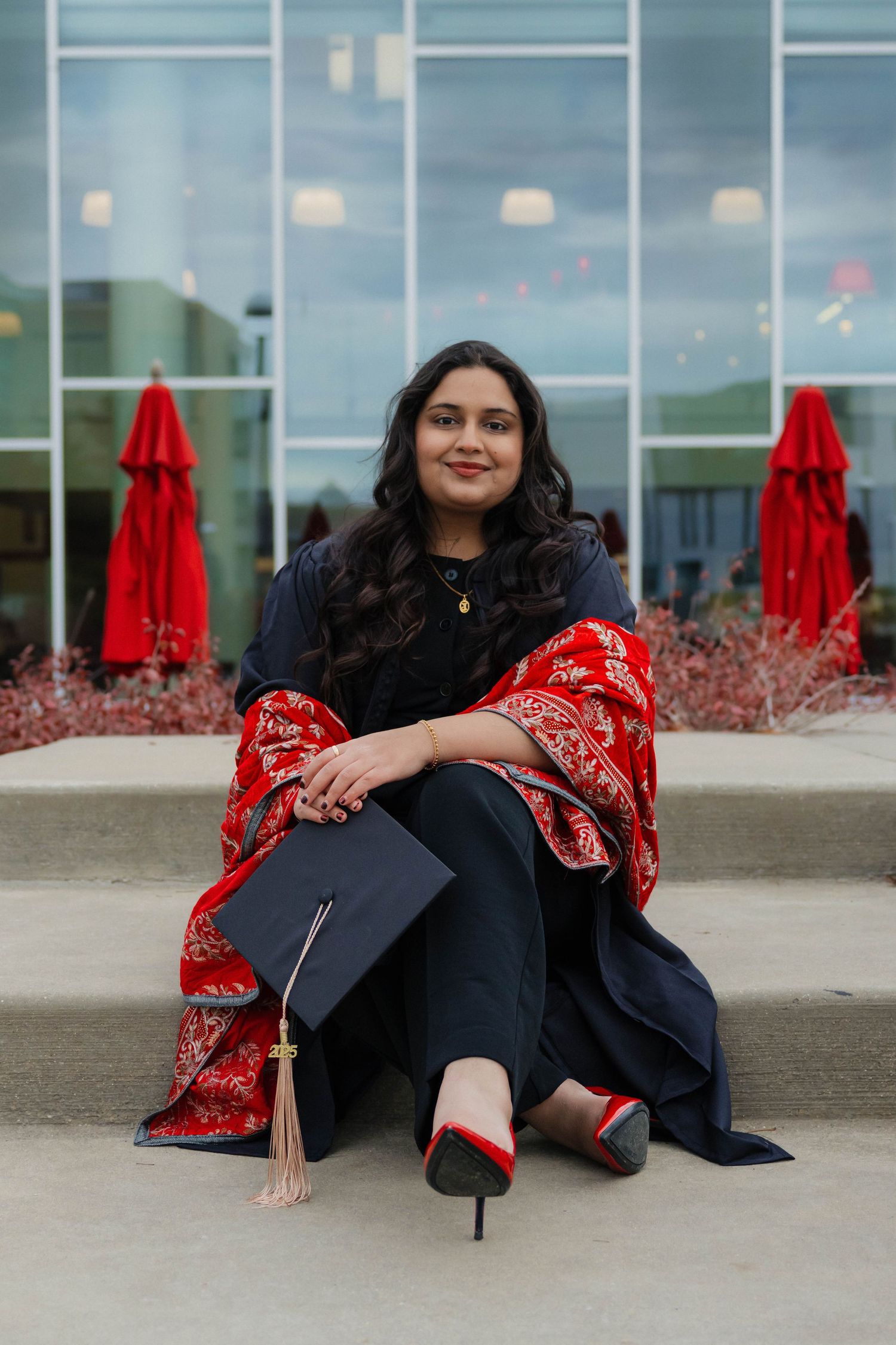Graduate in navy gown and red shawl sitting on steps holding a mortarboard cap outdoors.