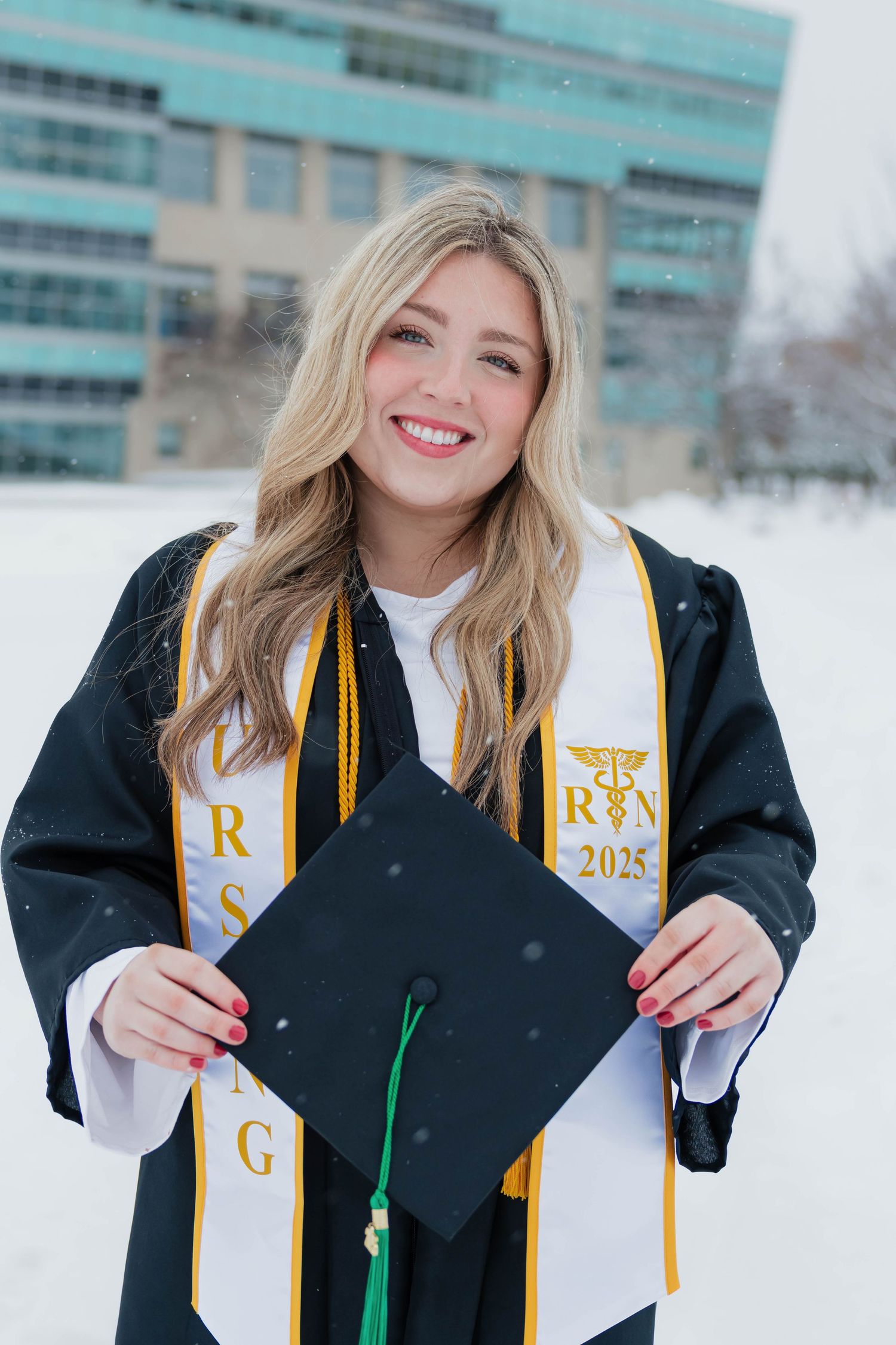 Blonde nursing graduate proudly displaying her mortarboard with RN 2025 stole in snowy campus setting.