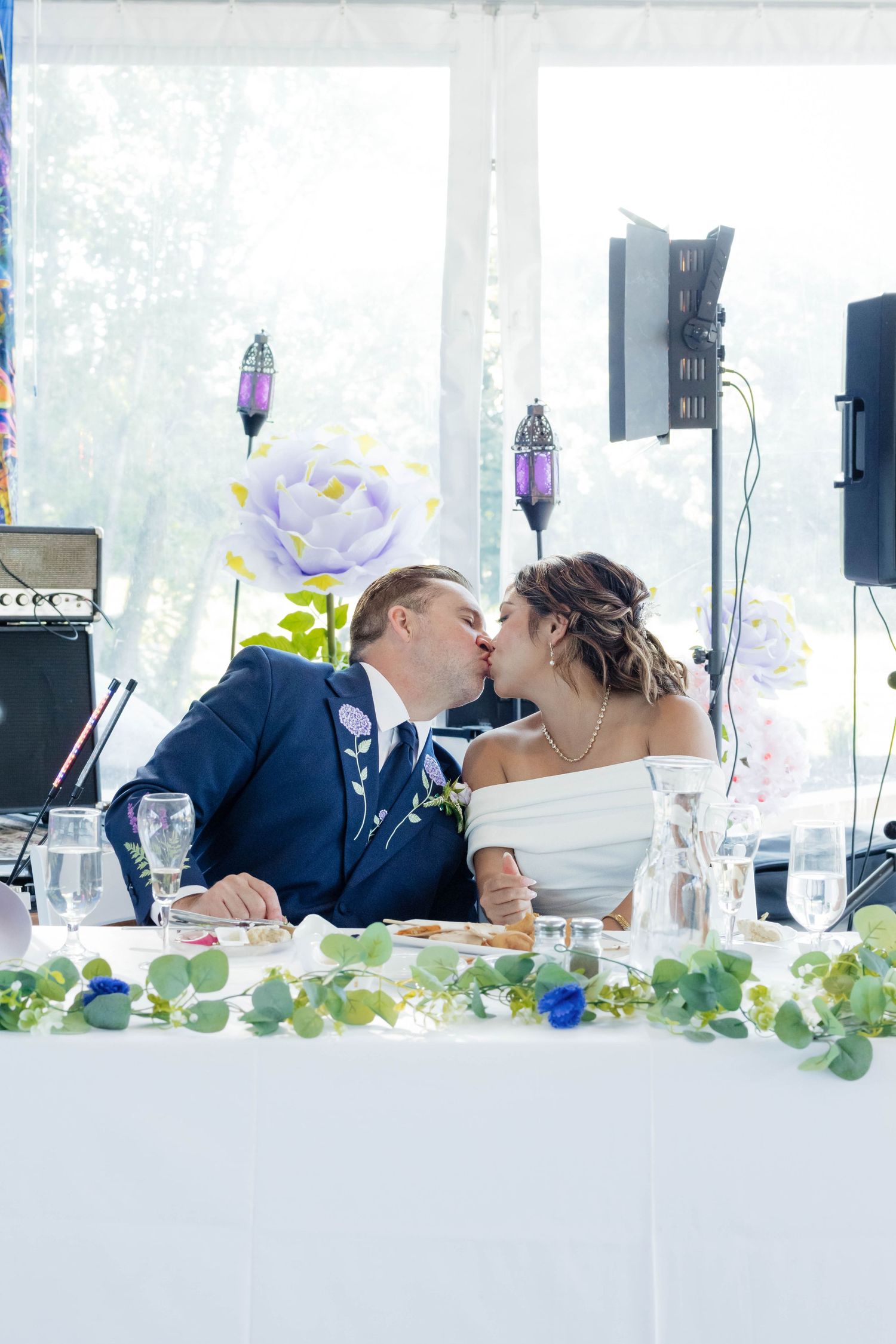 Bride and groom share a kiss at the sweetheart table during their wedding reception under a tent.