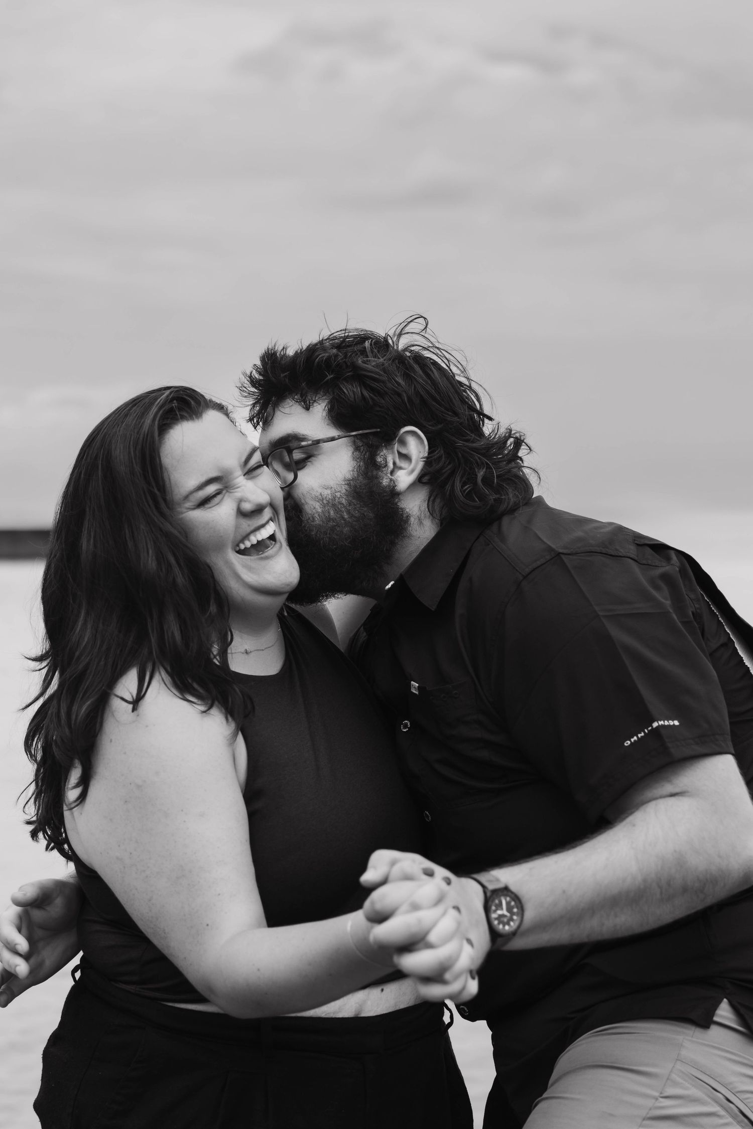 A black and white photo of a laughing couple holding hands as the man kisses the woman's cheek.