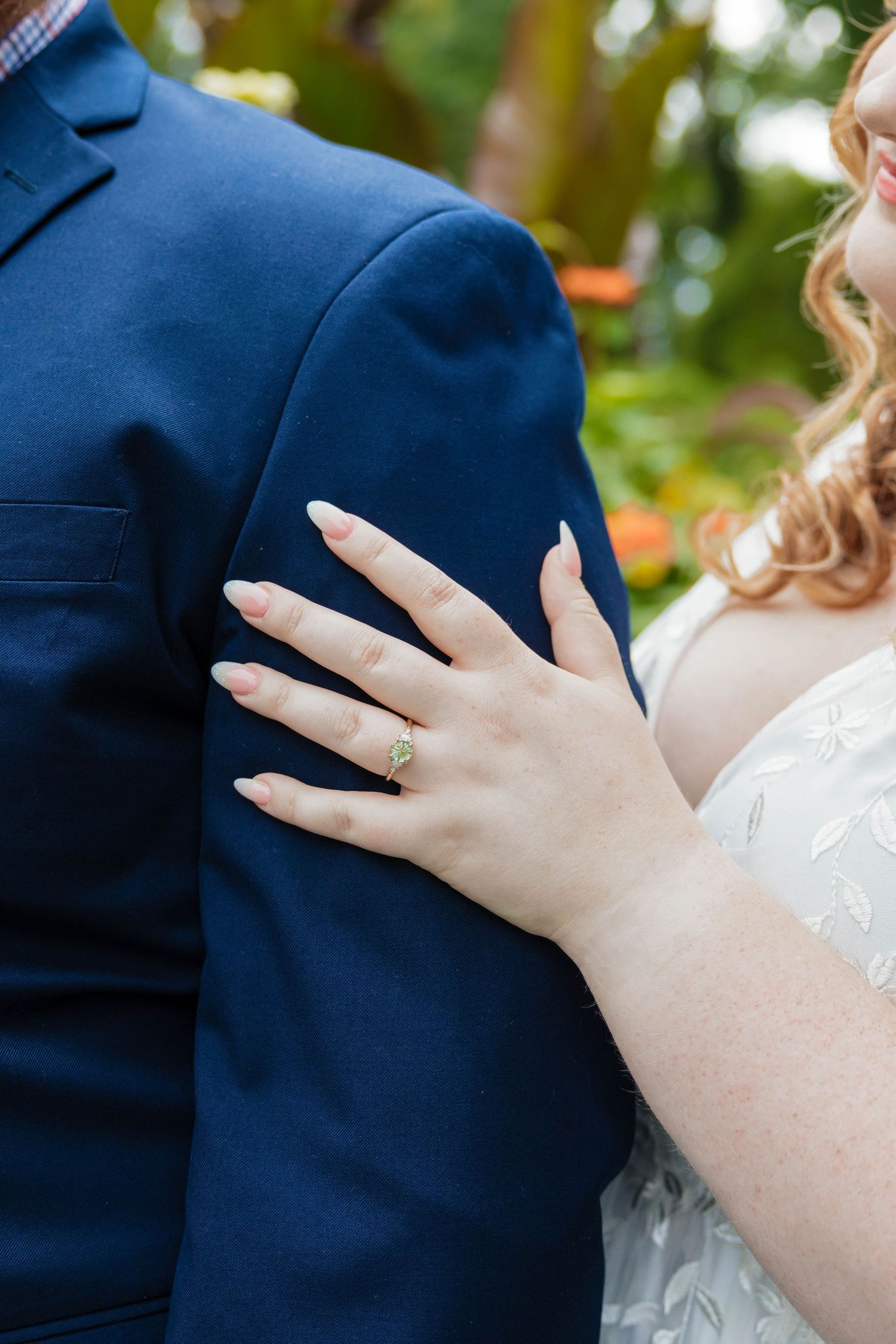 Close-up of bride's hand with a green gemstone engagement ring resting on groom's navy suit.
