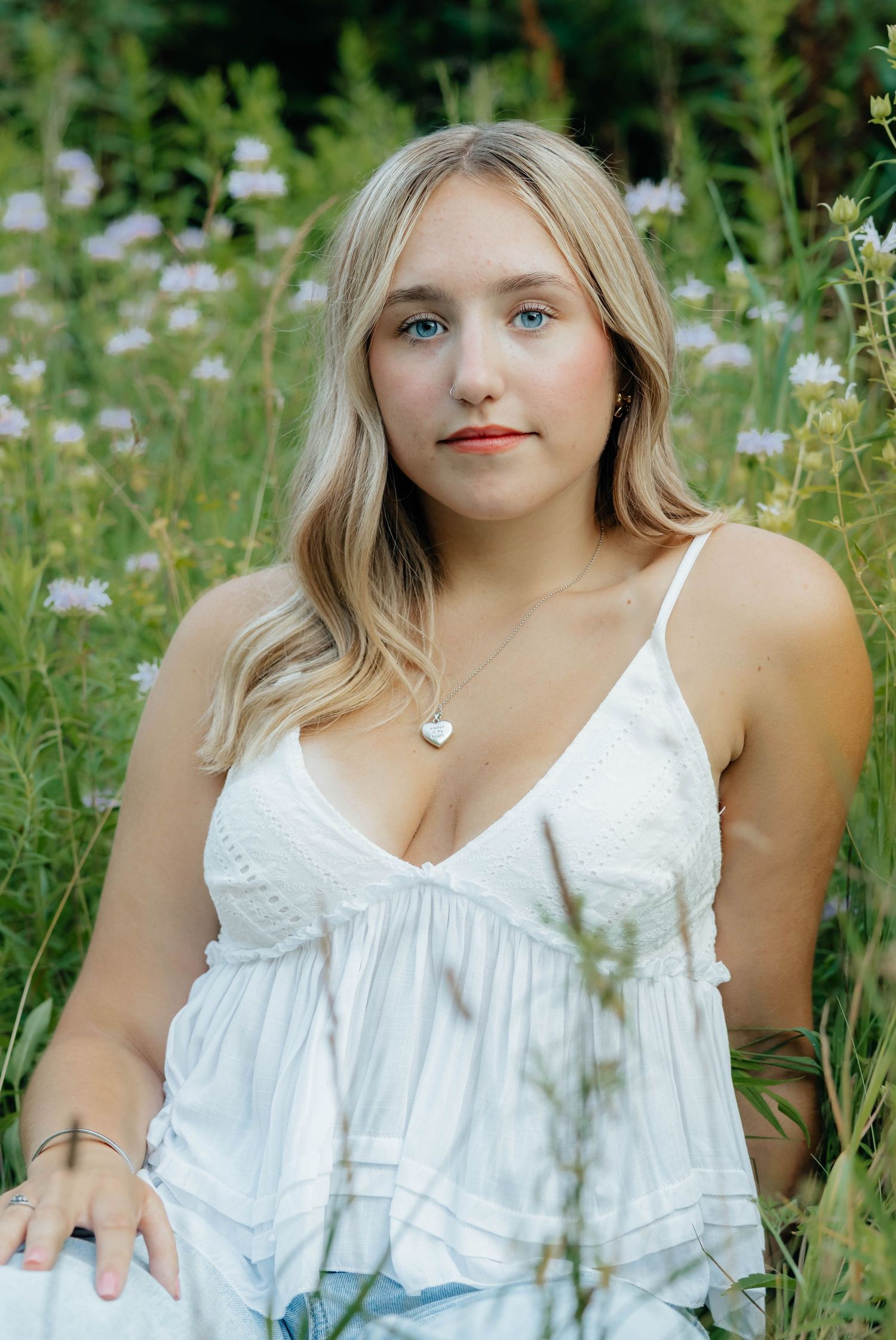 Blonde girl in white sundress with heart necklace sitting among wildflowers for senior portrait.