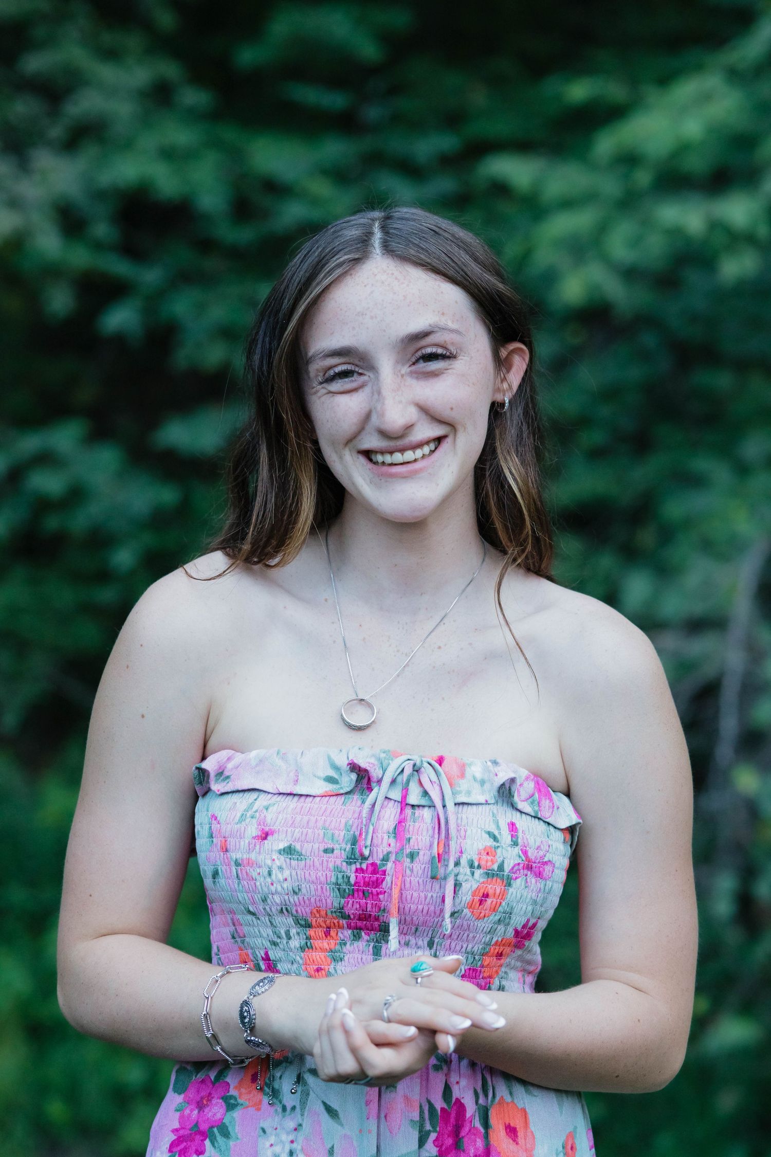 Smiling young woman in strapless floral dress wearing delicate necklace with green trees behind her.