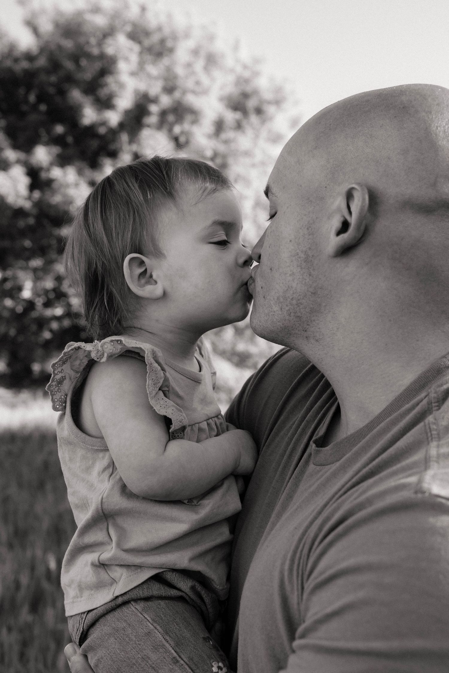 Black and white photo of bald father sharing sweet kiss with baby daughter outdoors.