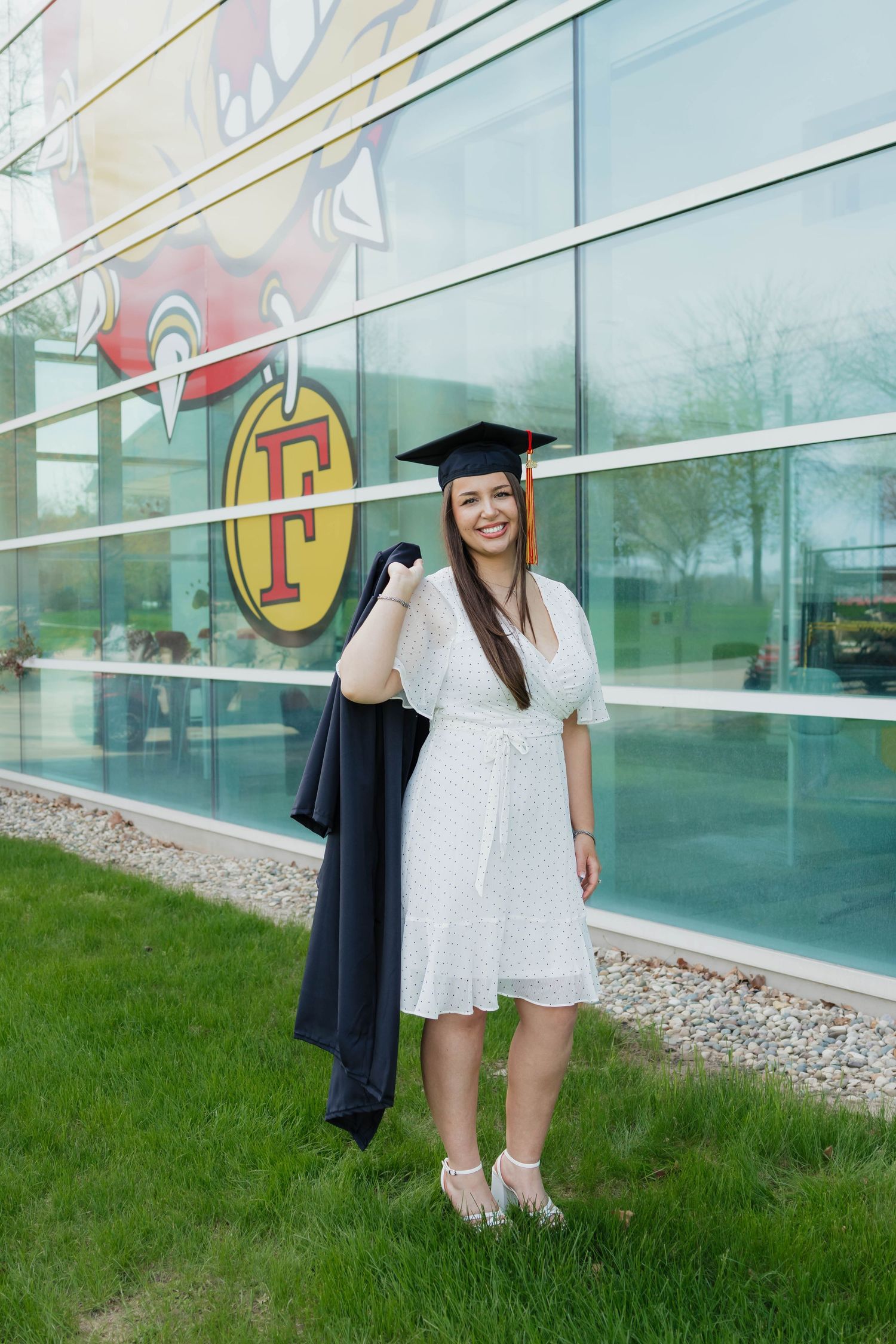 Female graduate in white dress holding black gown outside Ferris State University building.
