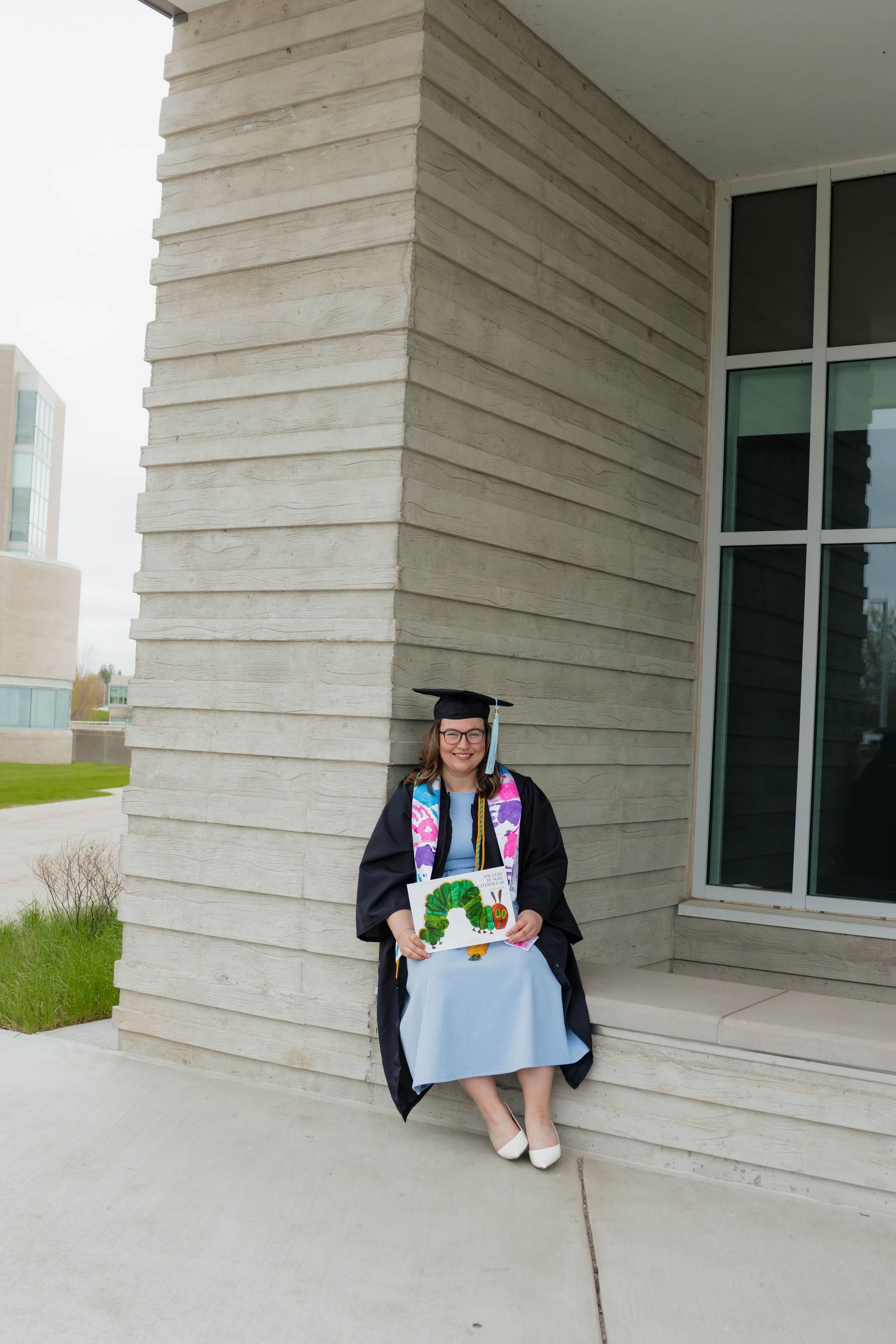 Graduate in cap and gown sitting on steps holding The Very Hungry Caterpillar book outside university building.