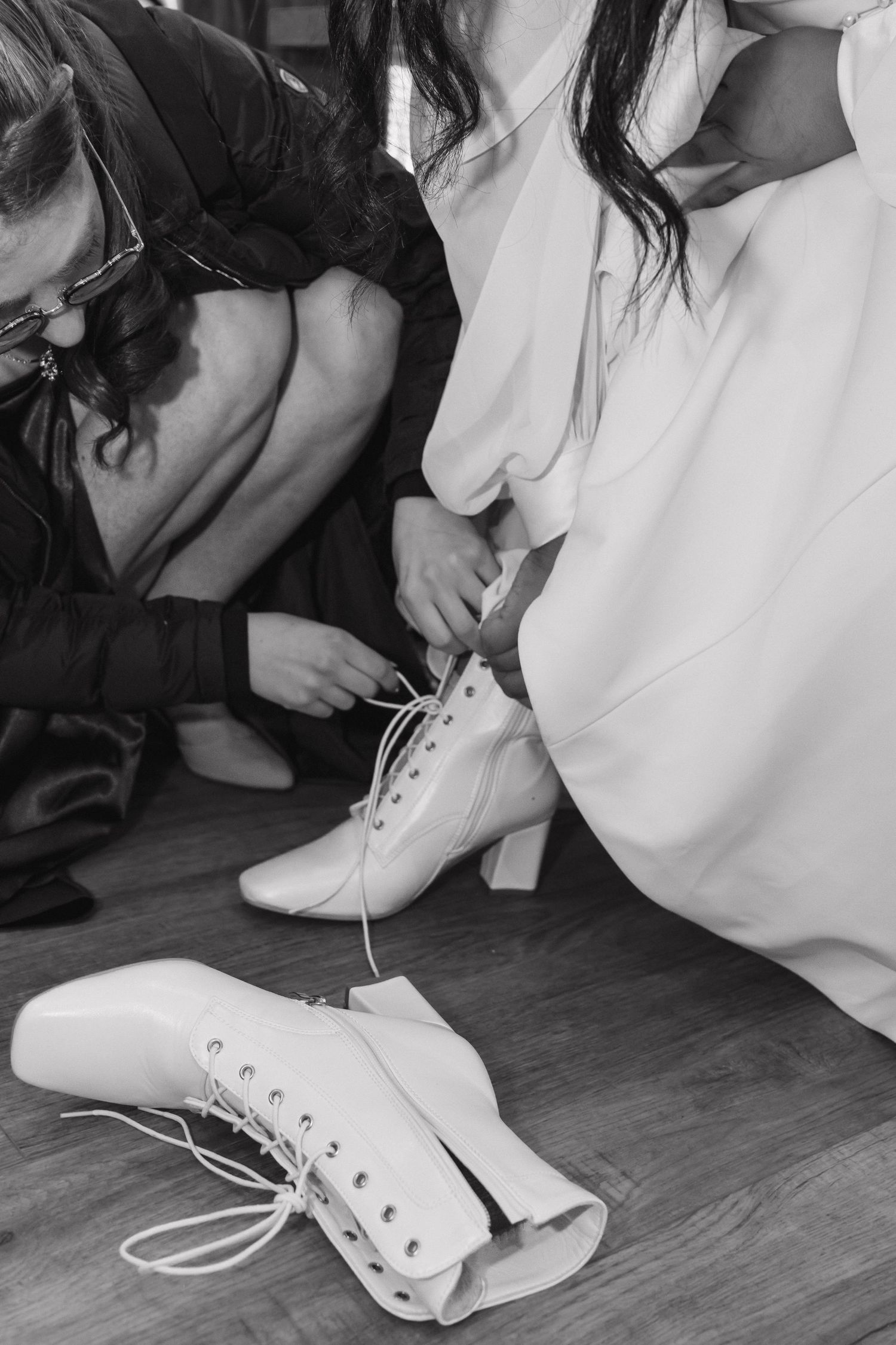 Black and white photo of bridesmaids helping bride lace up white heeled boots before wedding ceremony.