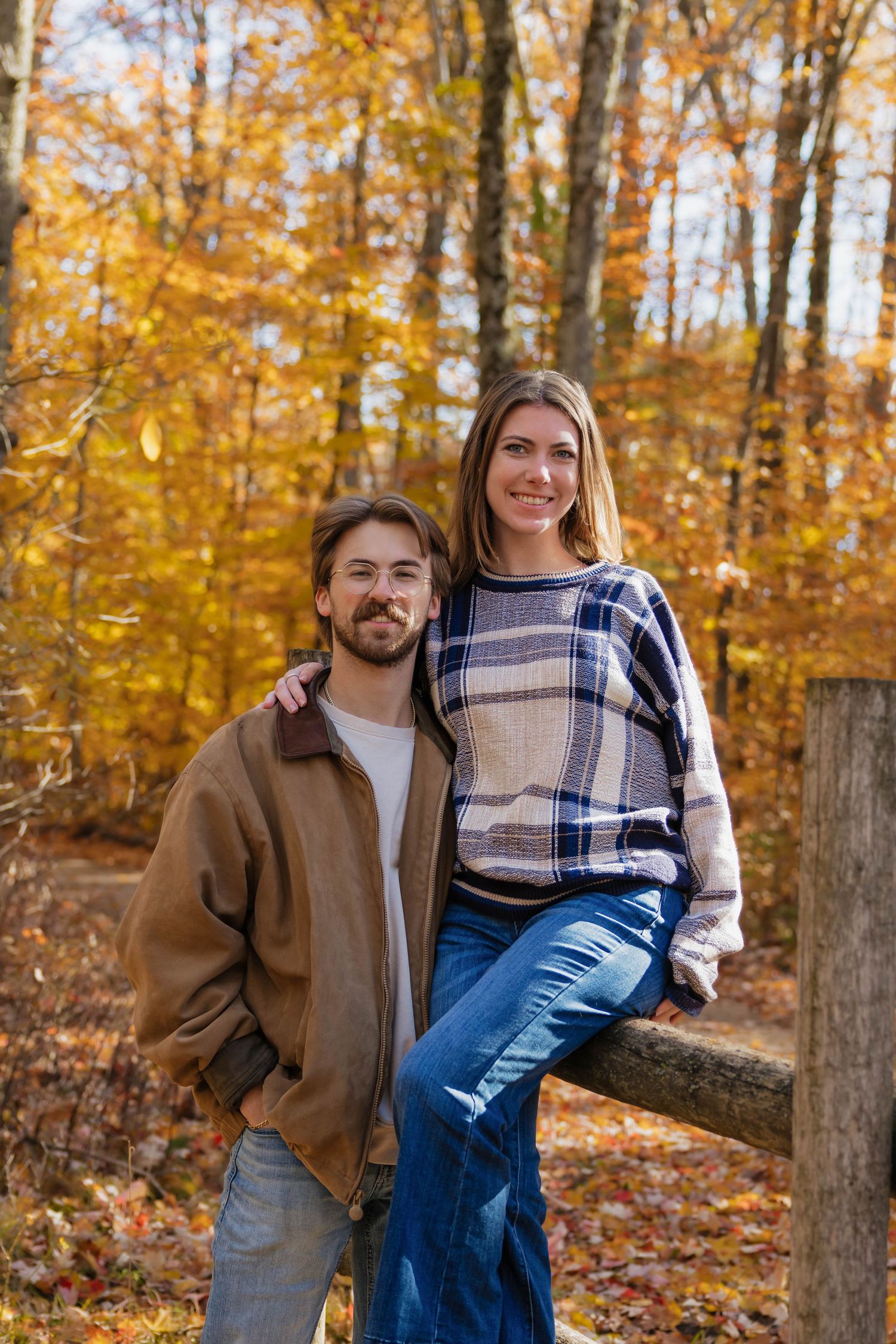 Couple smiling together in a vibrant autumn forest, leaning against a wooden fence rail.
