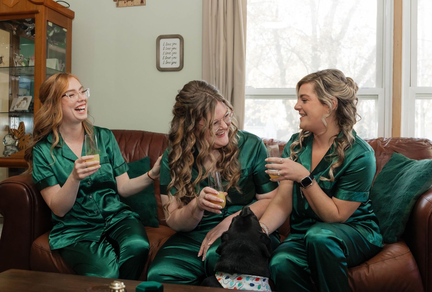Three bridesmaids in emerald green satin pajamas laugh together on brown leather couch during bridal prep.