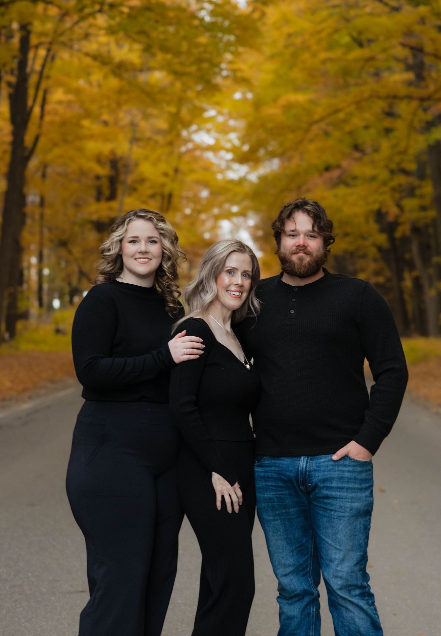 Three siblings in black outfits posing together on road with stunning yellow autumn trees.