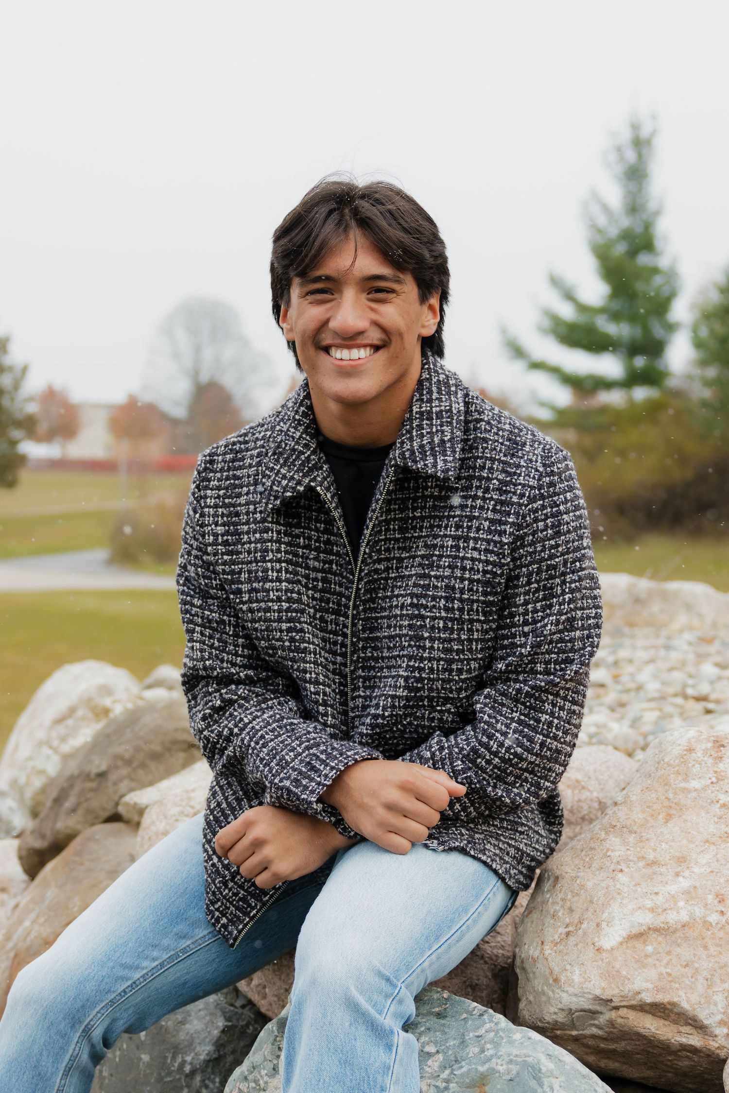 Smiling young man in tweed jacket sitting on rocks during autumn outdoor portrait session.