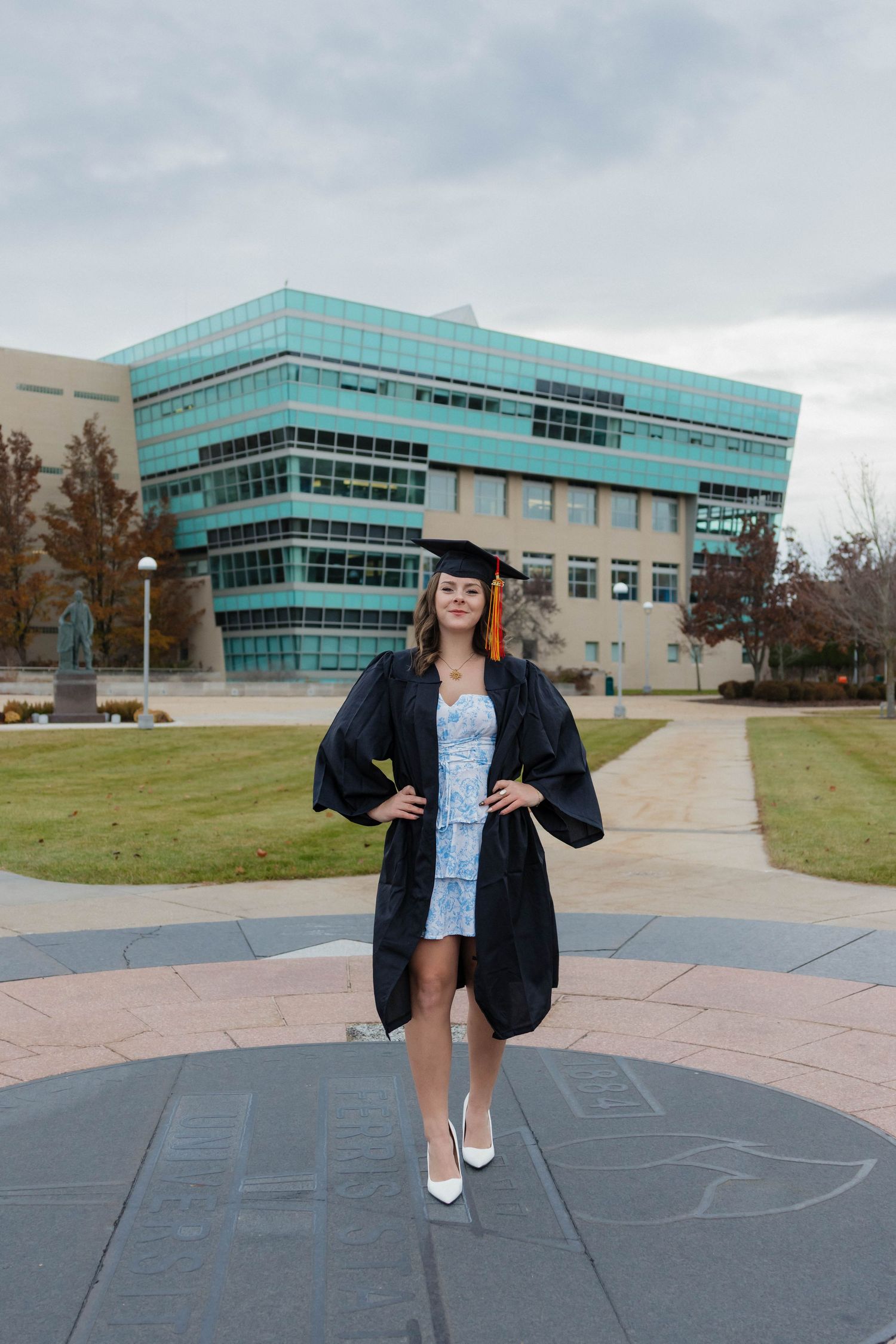 Female graduate in black cap and gown standing confidently on campus plaza with modern building behind her.