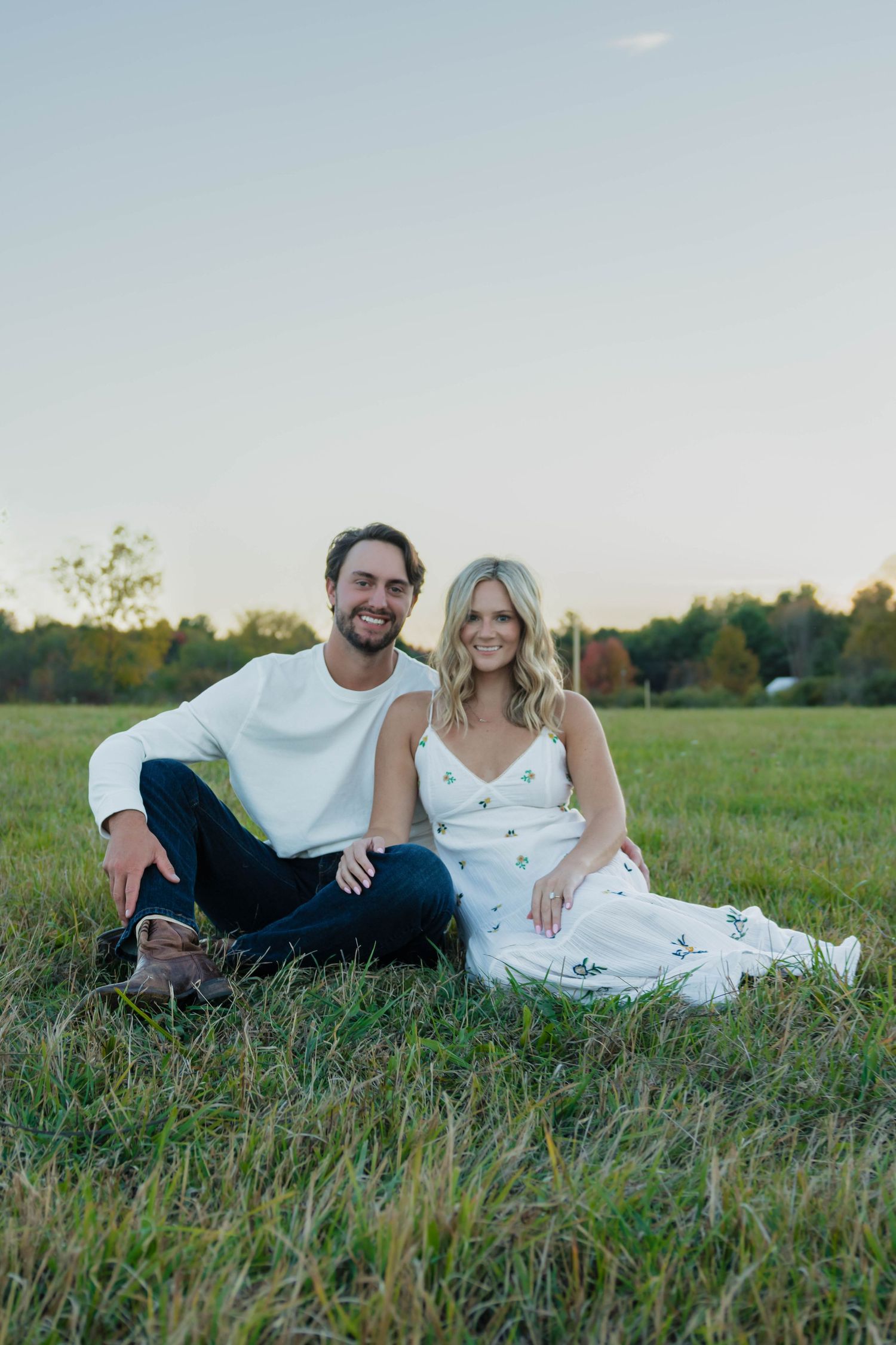 Happy engaged couple smiling and sitting together in a green field during a sunset engagement session.
