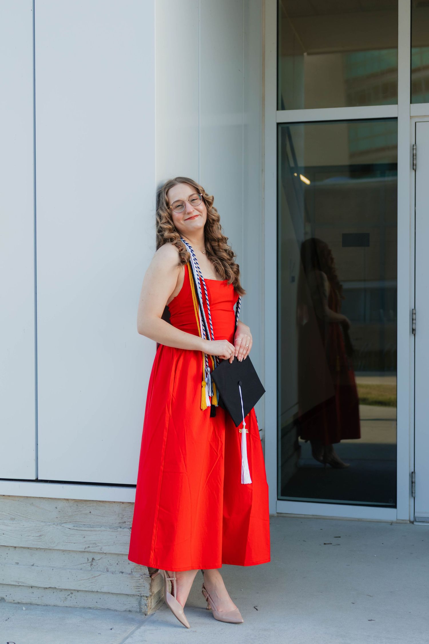 A smiling graduate in a red dress holds her cap while leaning against a white building exterior.
