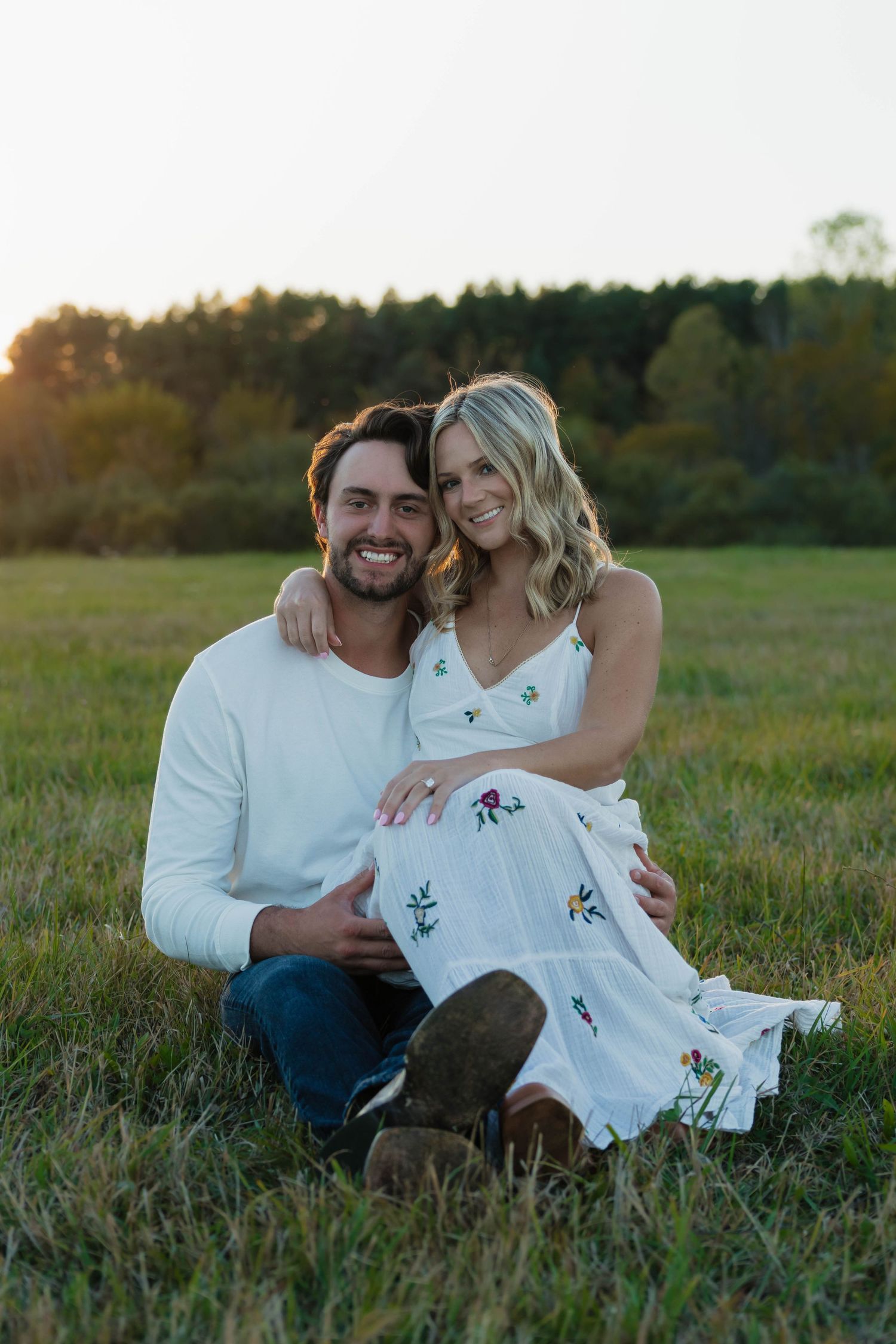 Smiling couple sitting together in a golden field at sunset during their engagement session.