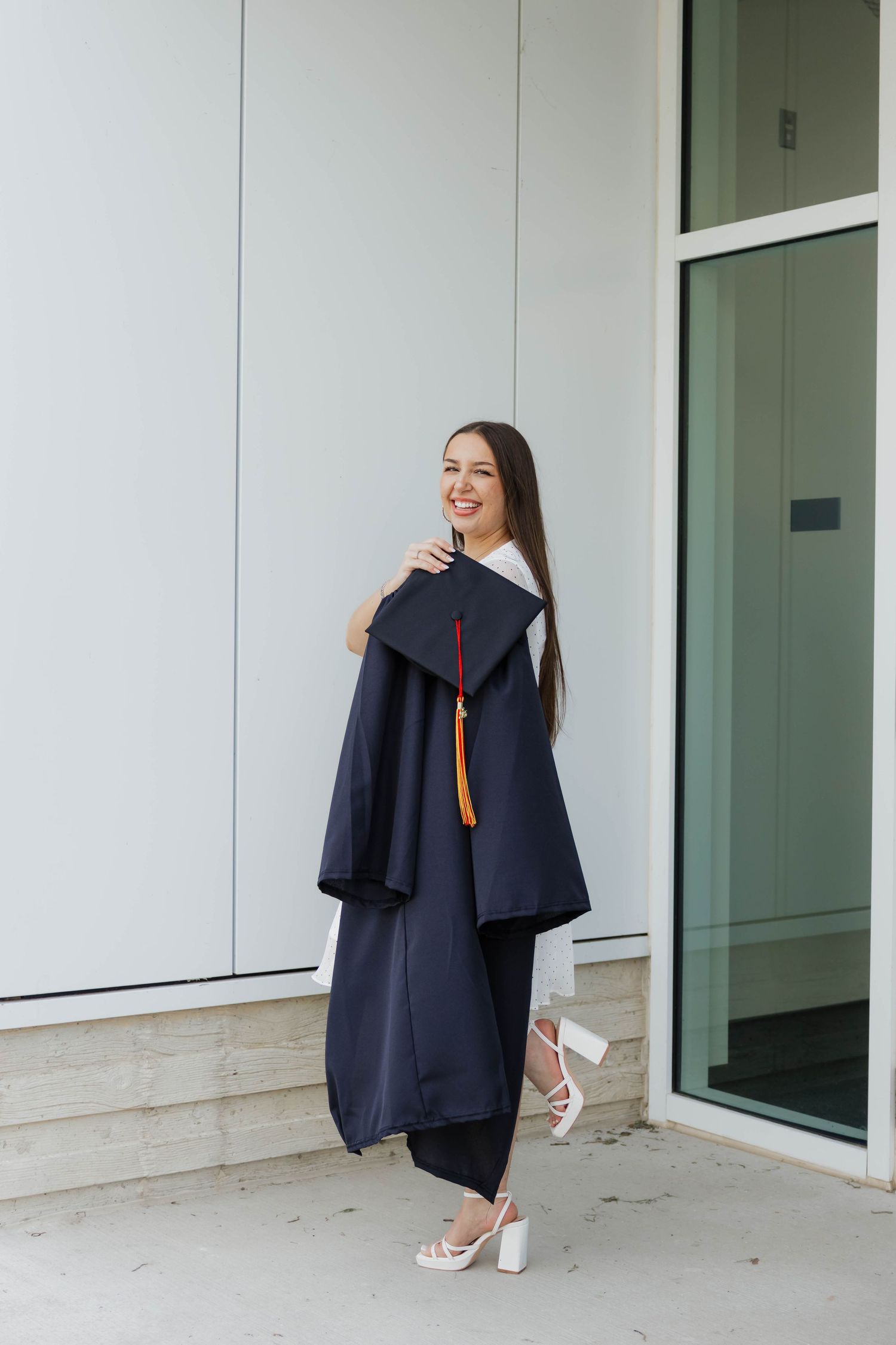 Smiling graduate holding navy cap and gown outside a modern building in heels.