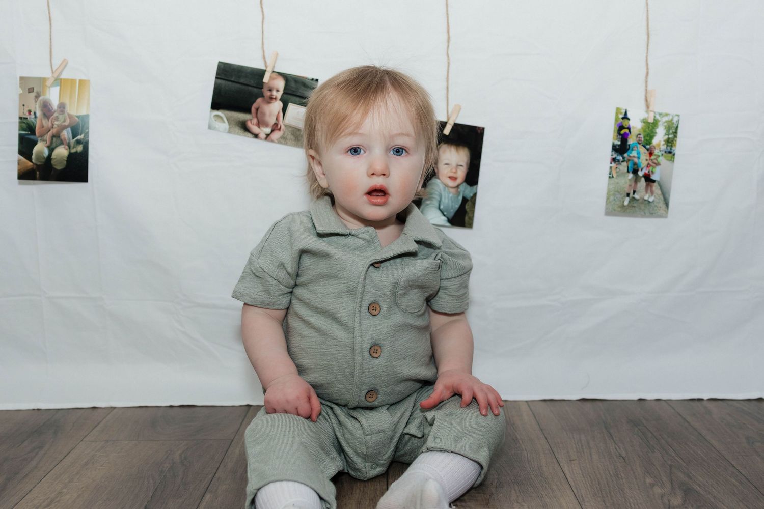 Adorable blue-eyed baby in sage green romper sitting in front of a white backdrop with hanging photos.