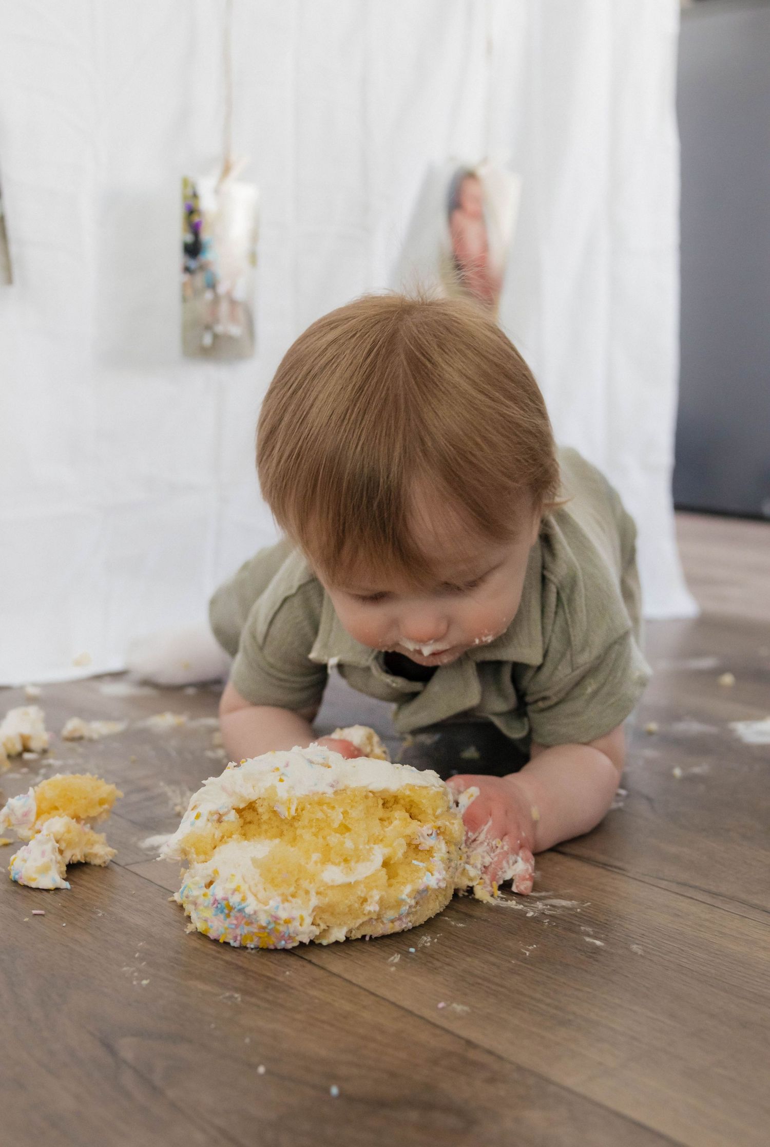 Baby in sage romper crawls toward smashed birthday cake on wood floor at cake smash.