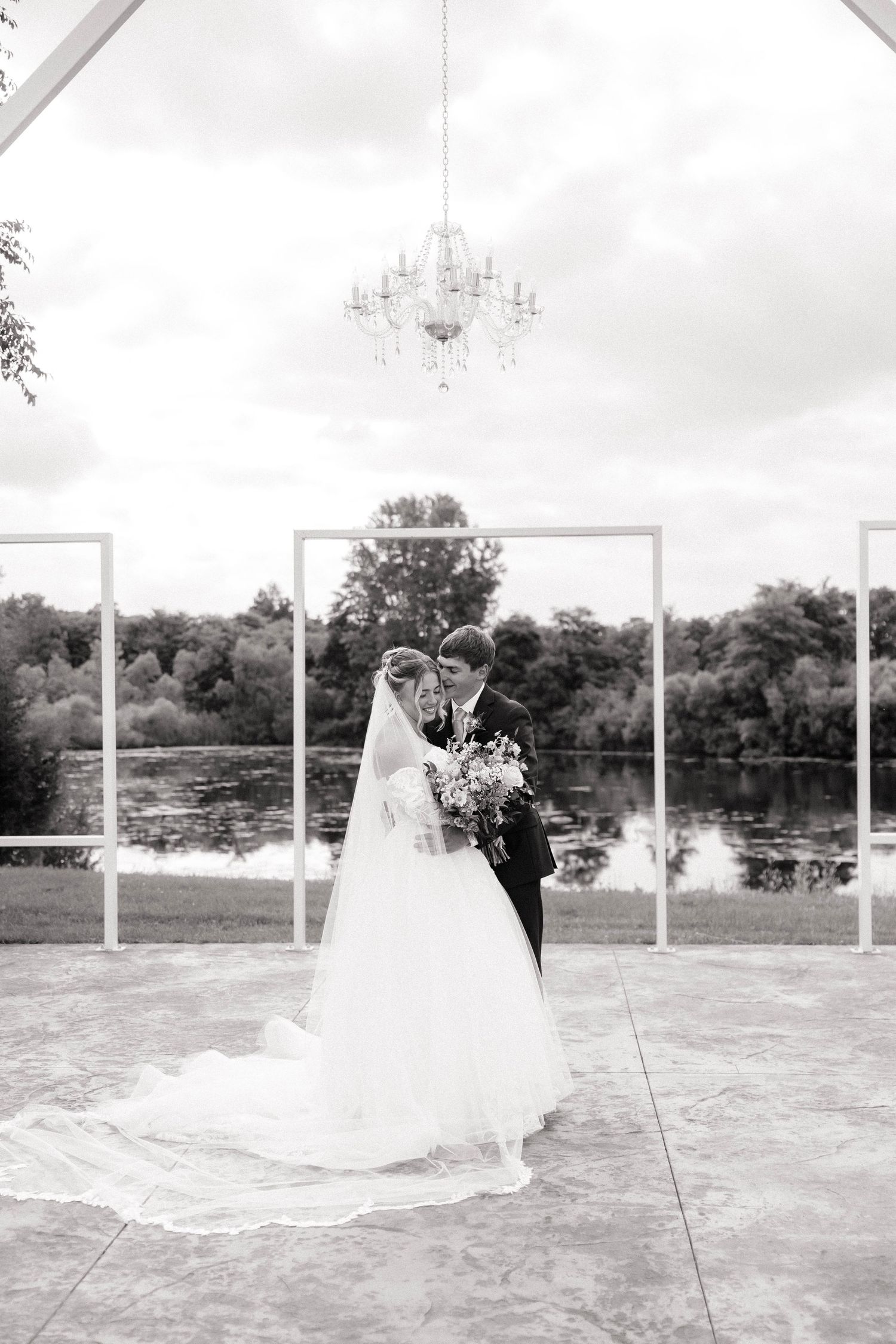 Black and white photo of bride and groom embracing under chandelier with lake backdrop at elegant outdoor wedding.