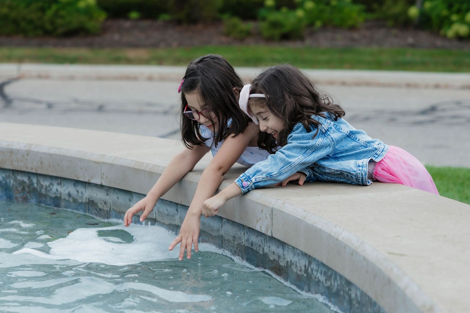 Two young girls leaning over fountain edge playing in water outdoors on sunny day.