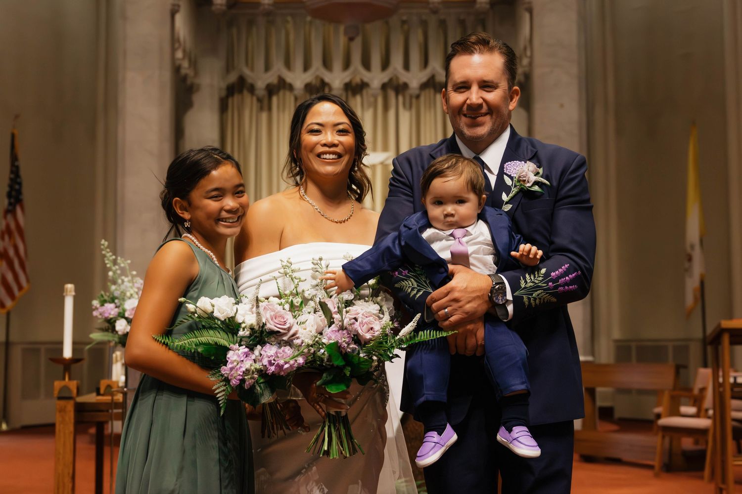Bride and groom smiling with a young bridesmaid and baby in a navy suit at church altar.