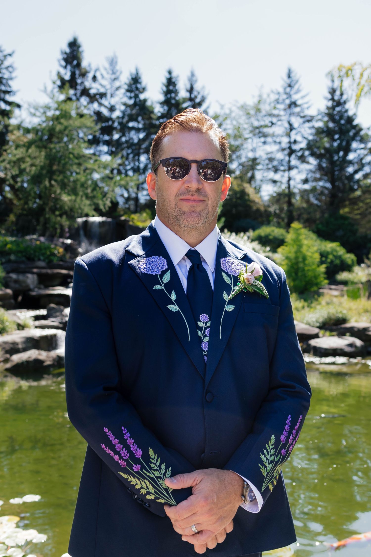 Dapper groom in navy embroidered suit with floral boutonniere stands confidently by garden pond wearing sunglasses.