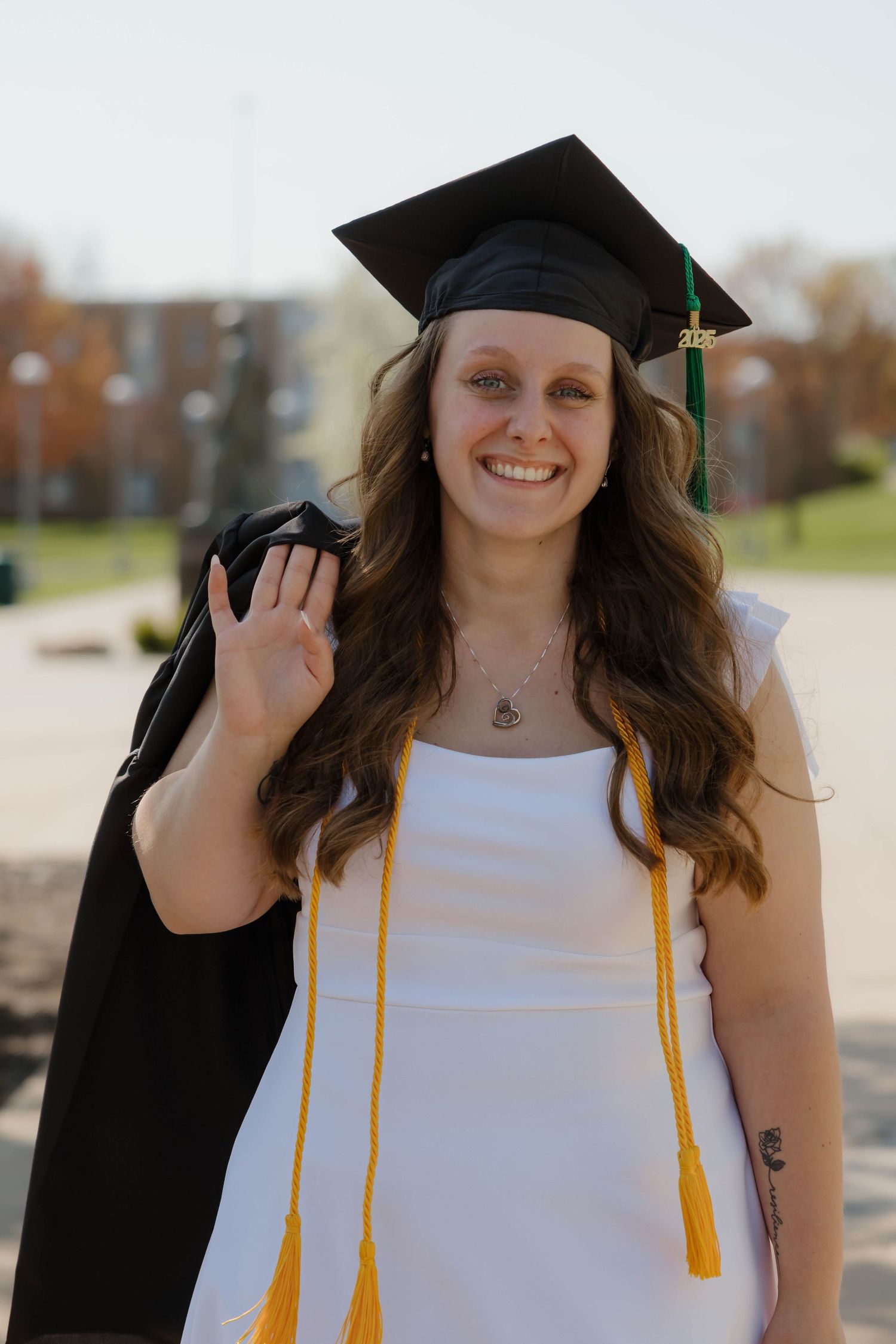 Smiling graduate in white dress and cap waving while holding black gown outdoors.