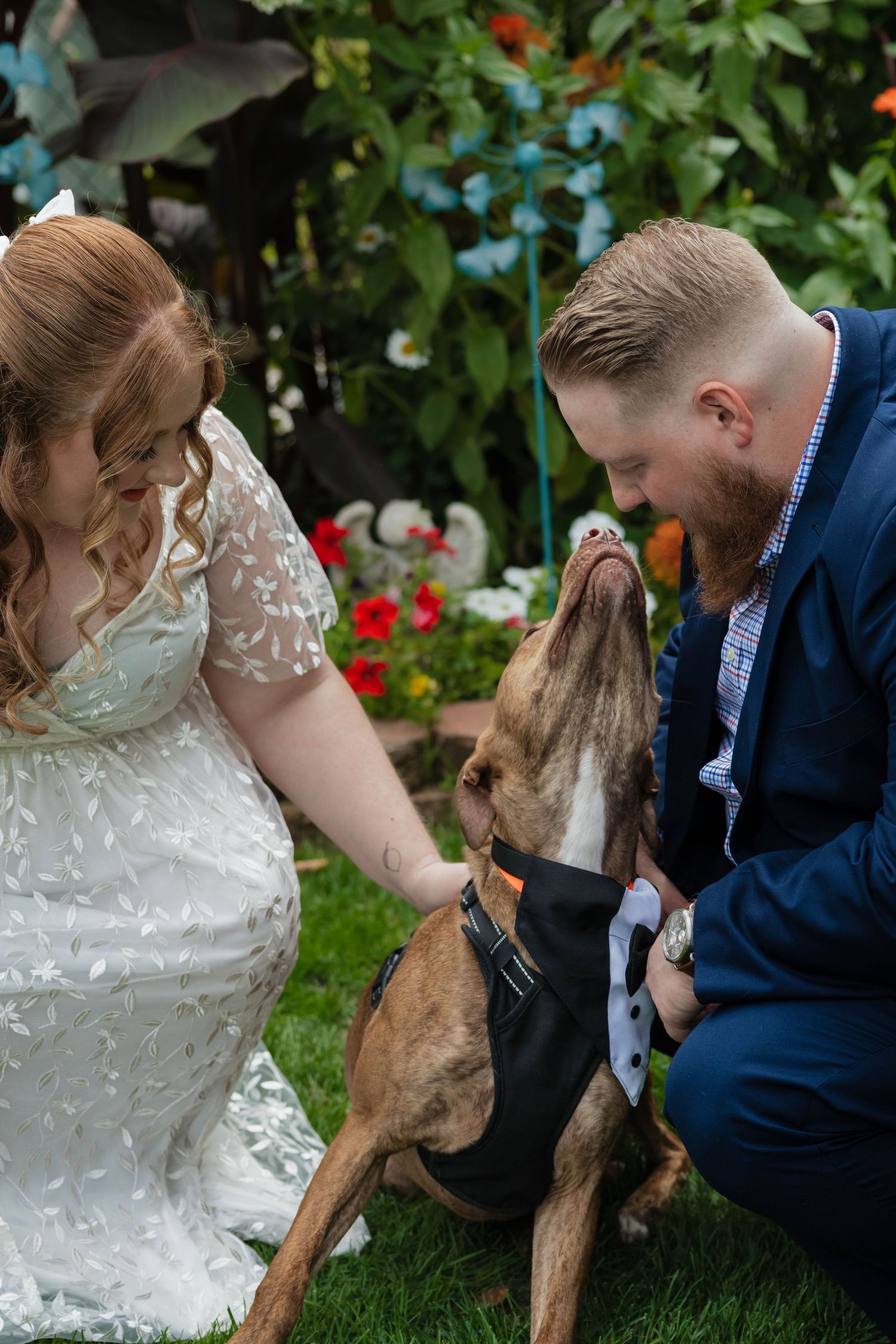 Wedding couple kneeling with brown dog in tuxedo bandana in colorful garden.