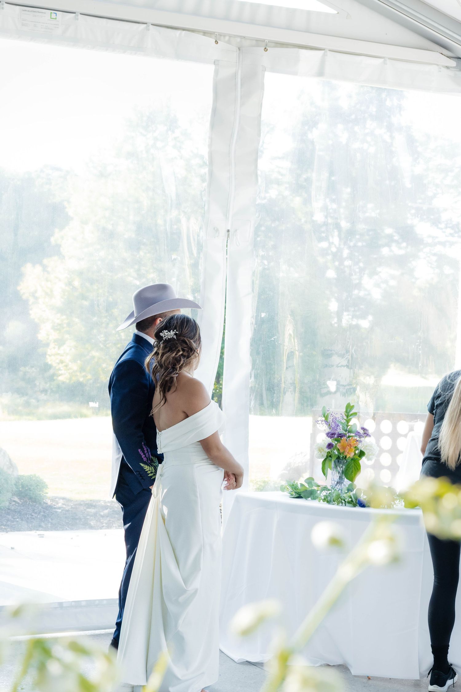 Bride and groom stand together at outdoor wedding ceremony altar beneath white tent with garden views.