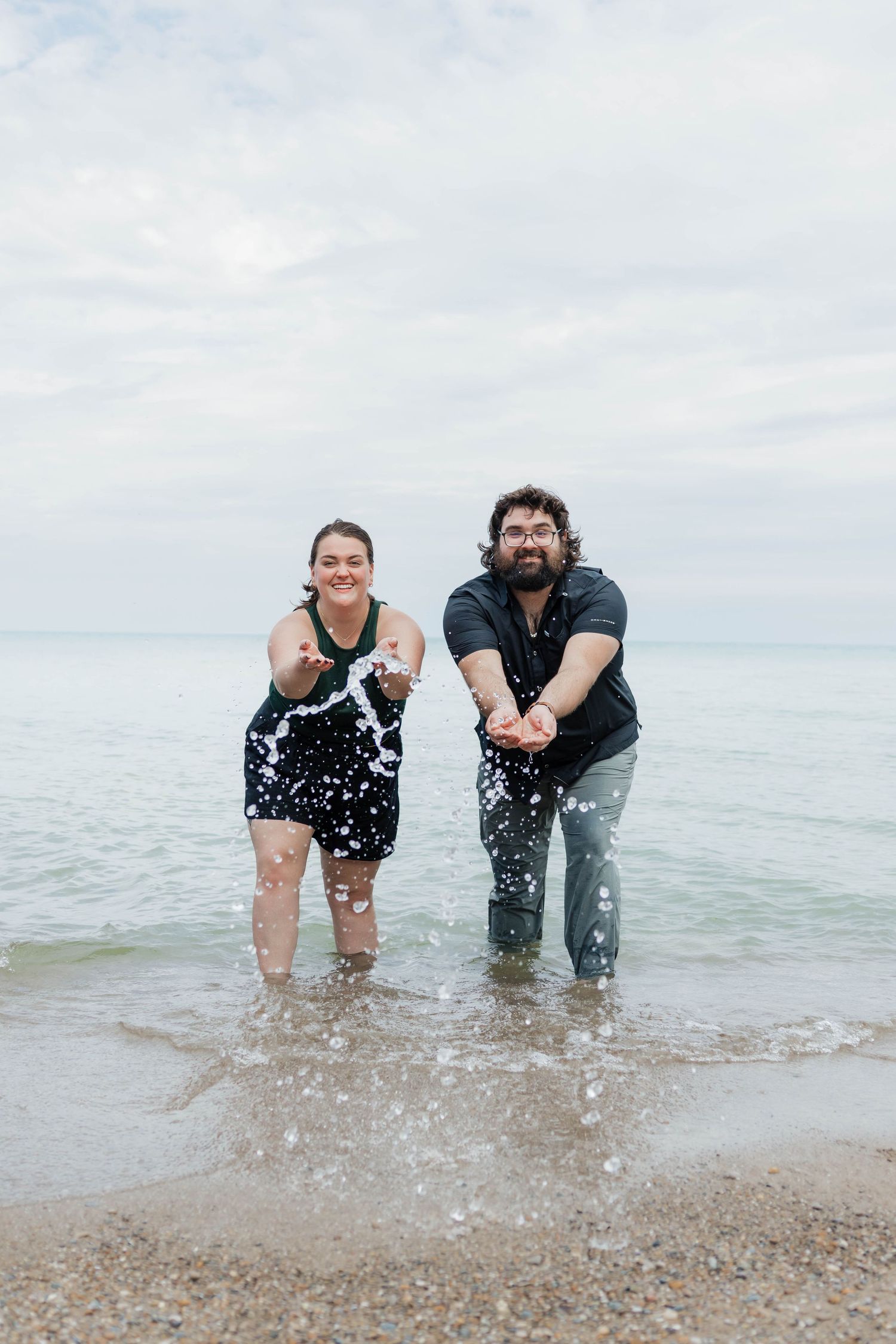 A happy couple stands in shallow ocean water, splashing and laughing together on a cloudy day.