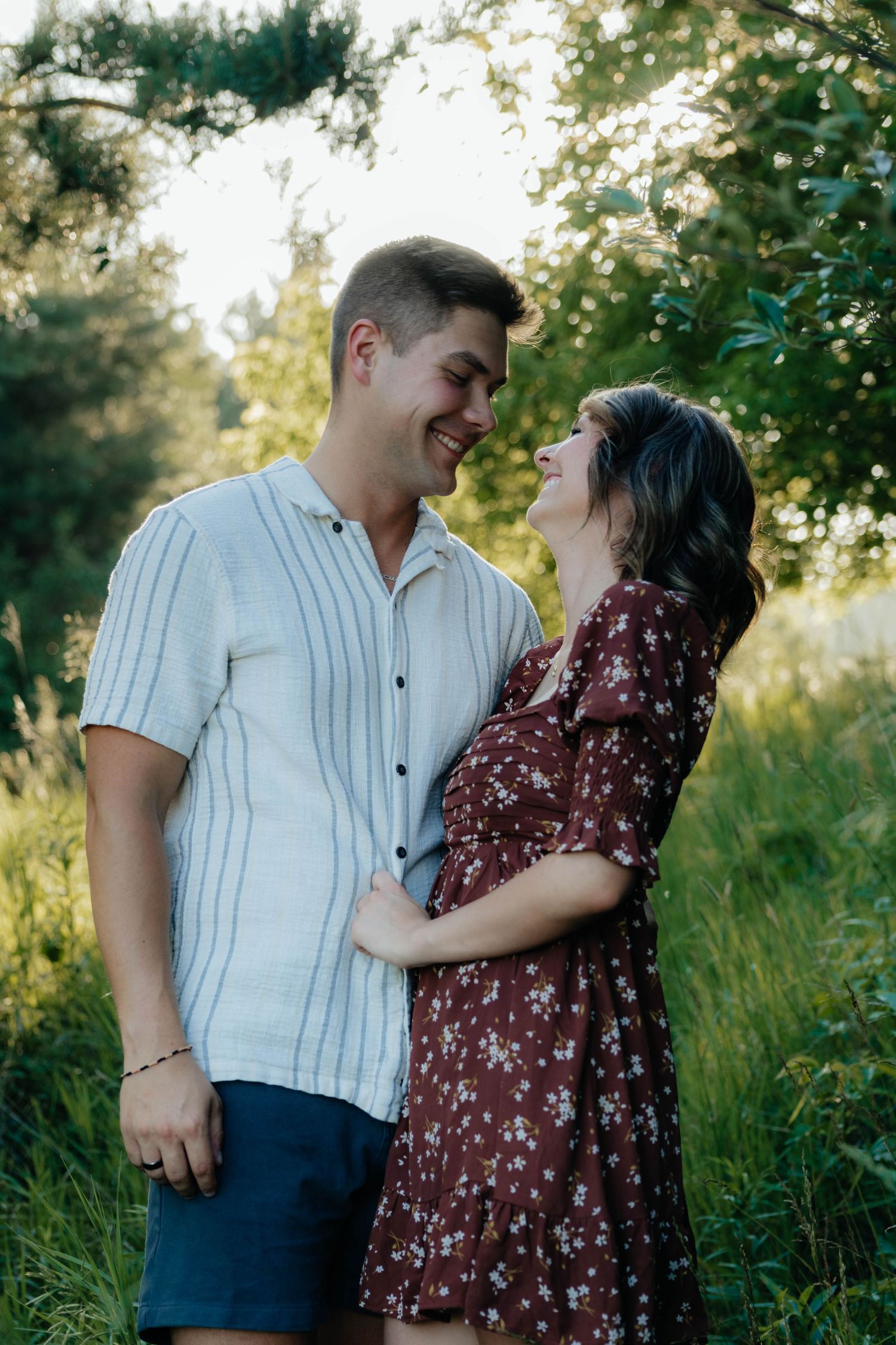 Happy couple embracing and laughing together outdoors surrounded by green trees and tall grass in warm sunlight.