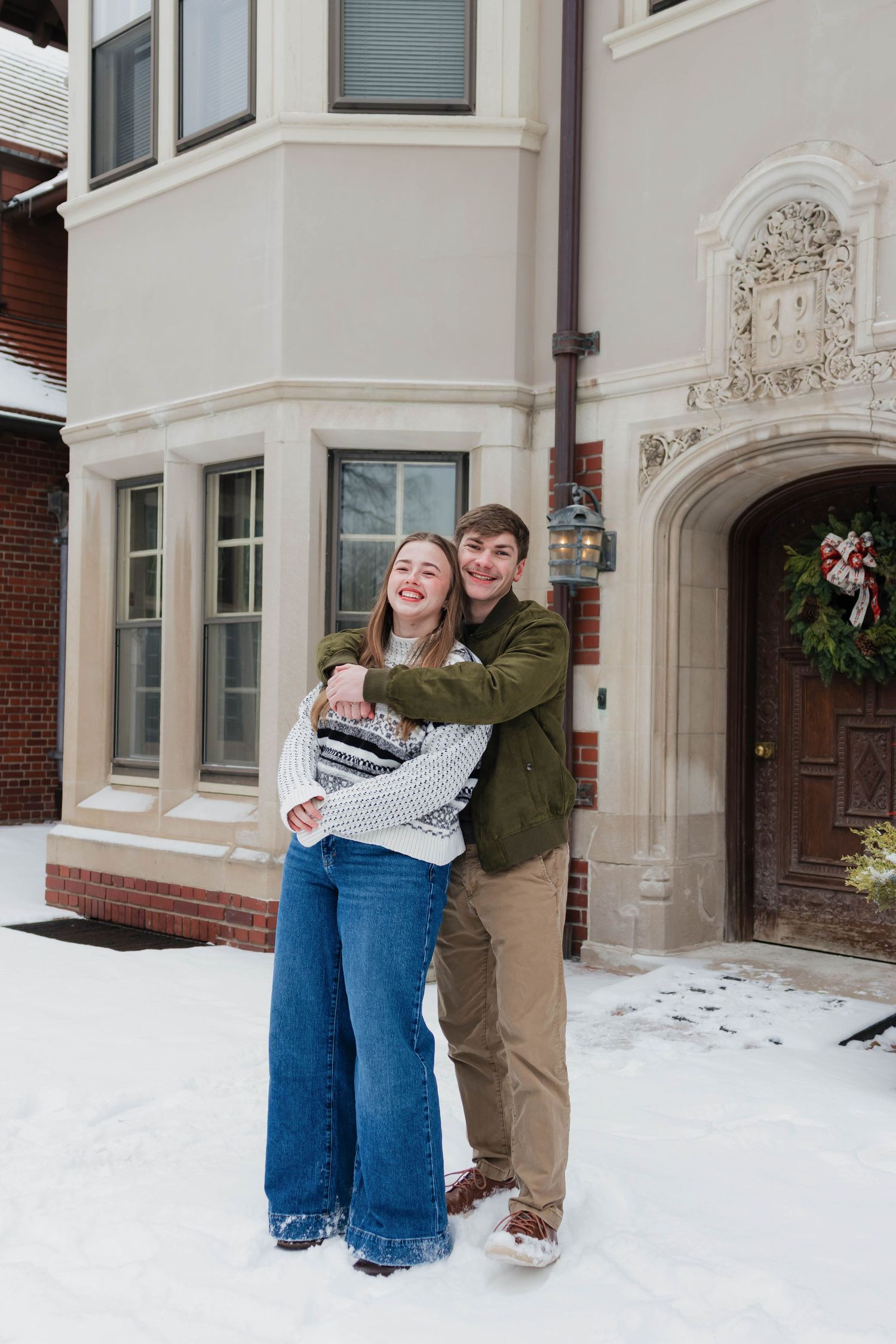 Happy couple embracing and smiling outside an elegant snow-covered building with holiday decorations.