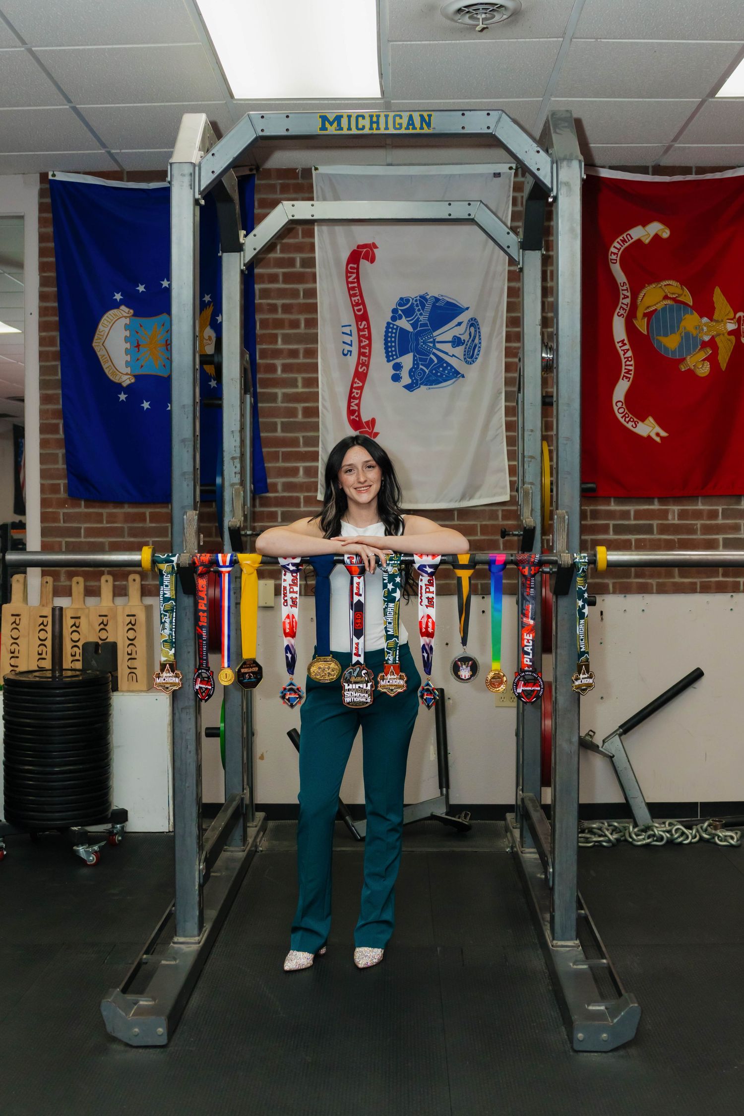Athlete posing with medals on power rack beneath military branch flags in Michigan weight room.
