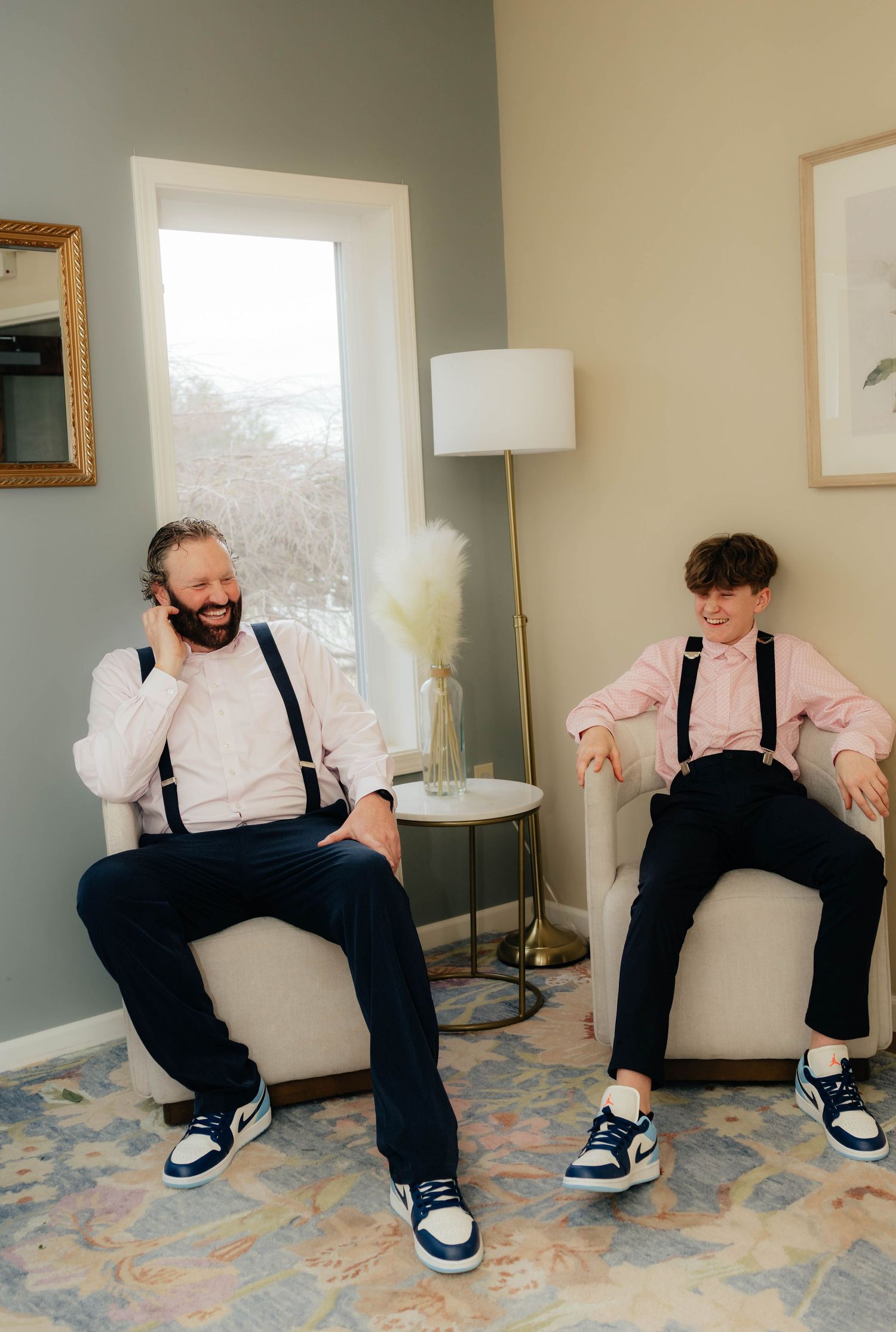 Groom and young boy in matching pink shirts and suspenders laughing together while seated in chairs.