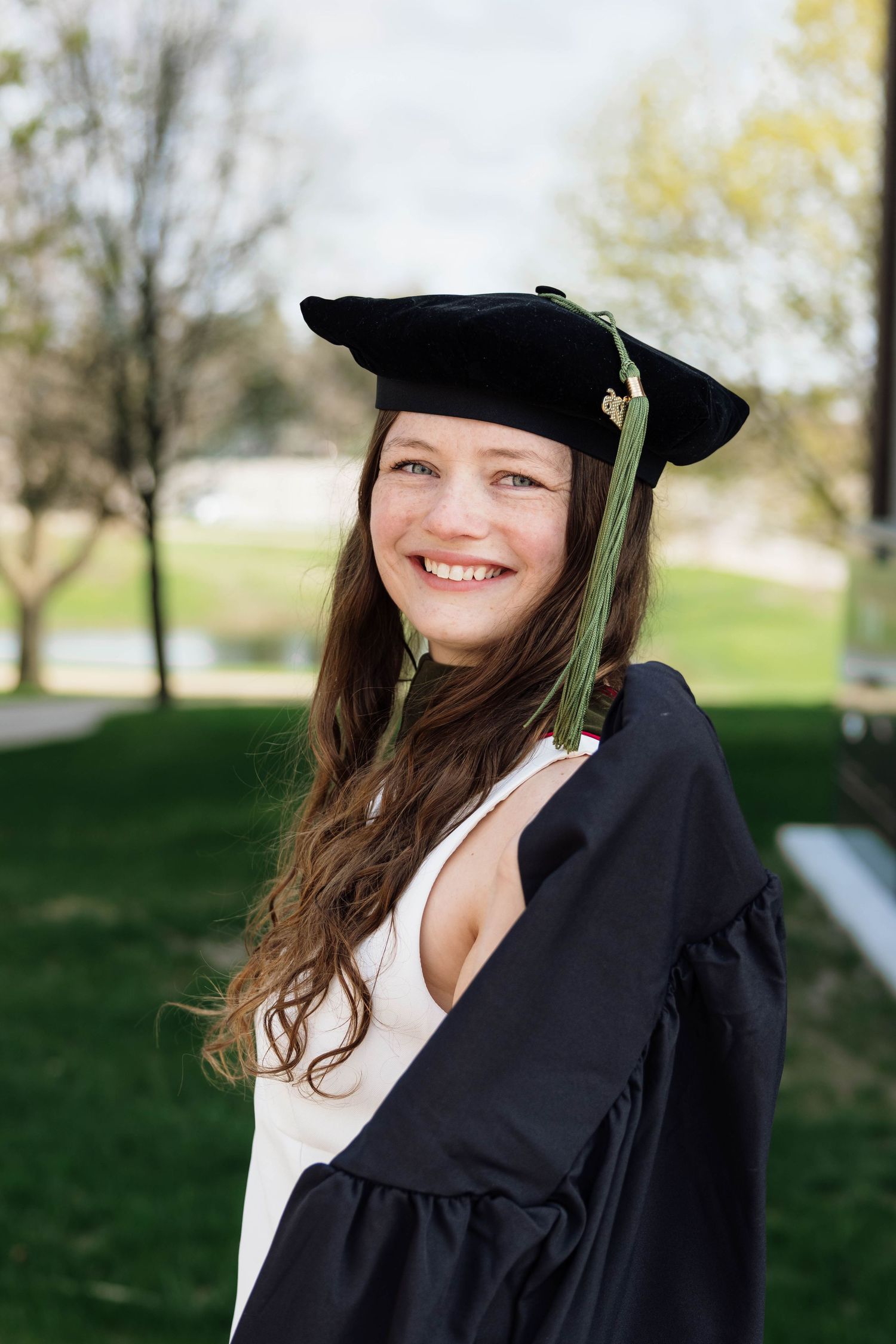 Smiling brunette graduate in black cap with green tassel and gown poses in park setting.