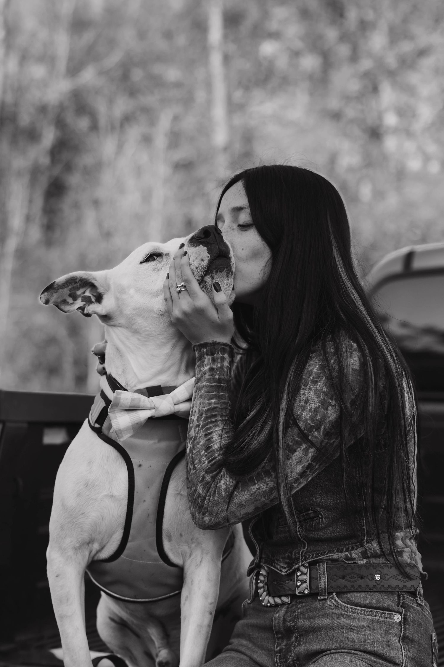 Black and white portrait of woman kissing a white pitbull dog on the nose outdoors.