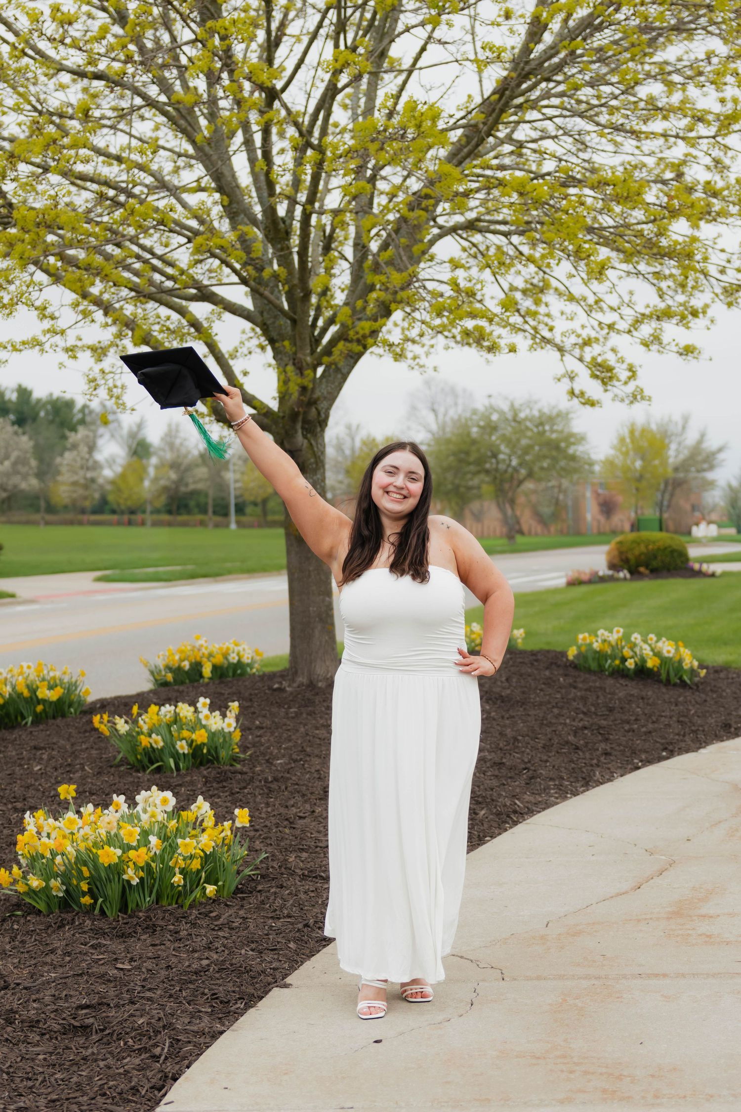 Smiling graduate in white dress holding mortarboard cap outdoors near spring flowers and blooming trees.