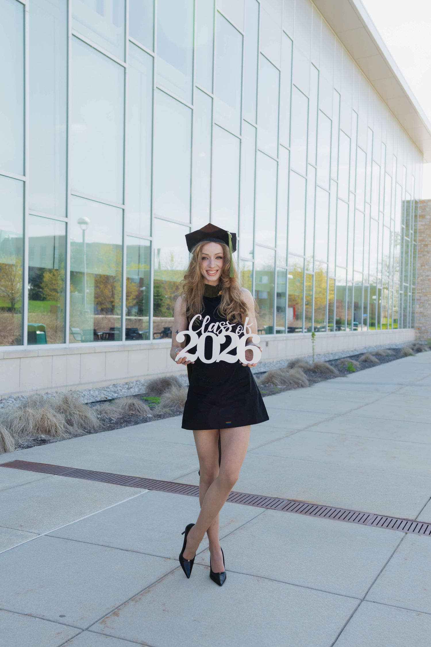 A smiling graduate in black dress and cap holds a Class of 2025 sign outside a modern building.